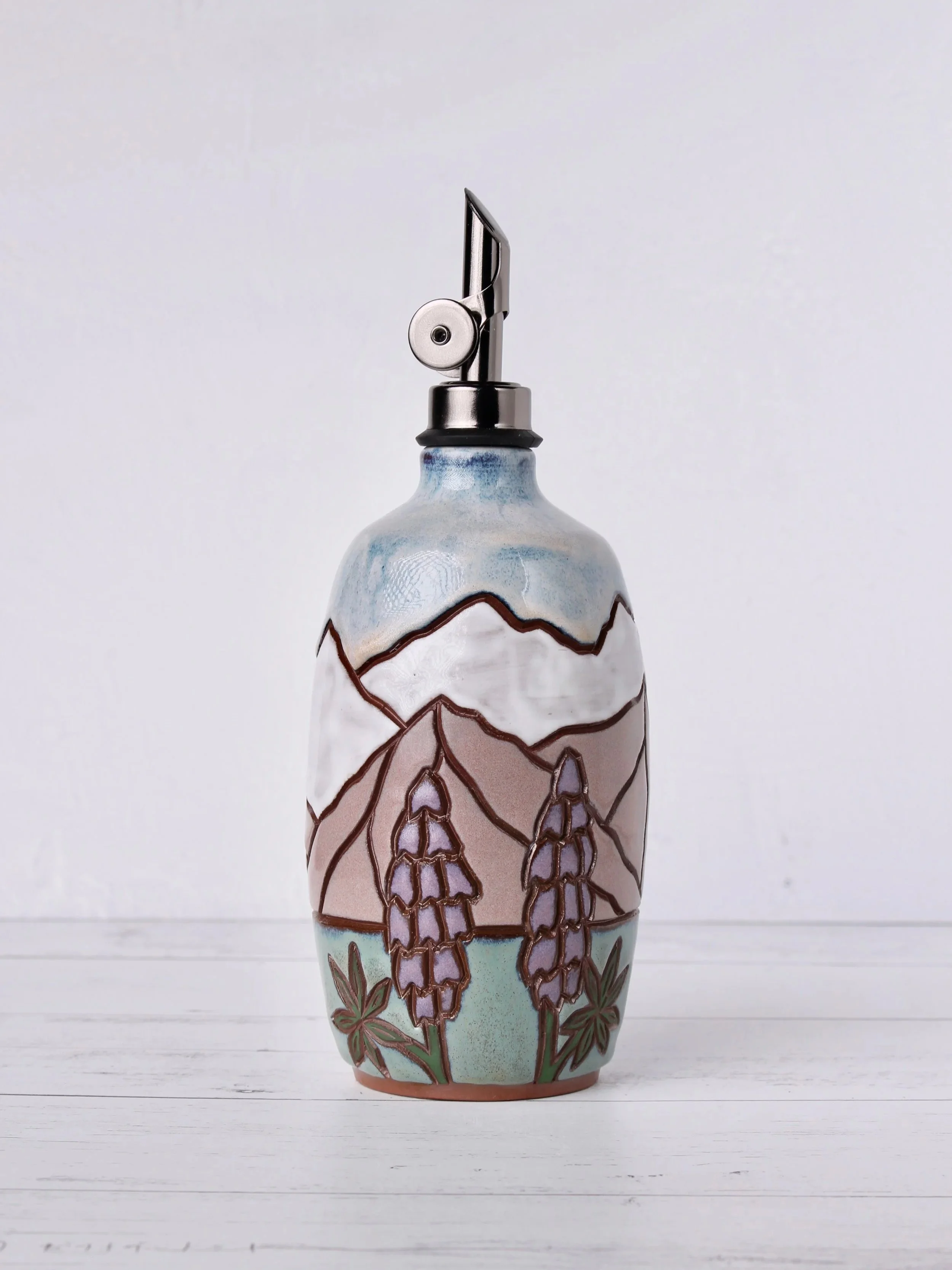 Decorative ceramic oil cruet with floral and mountain patterns, painted in soft pastel colors, sitting on a white wooden surface with a plain white background.