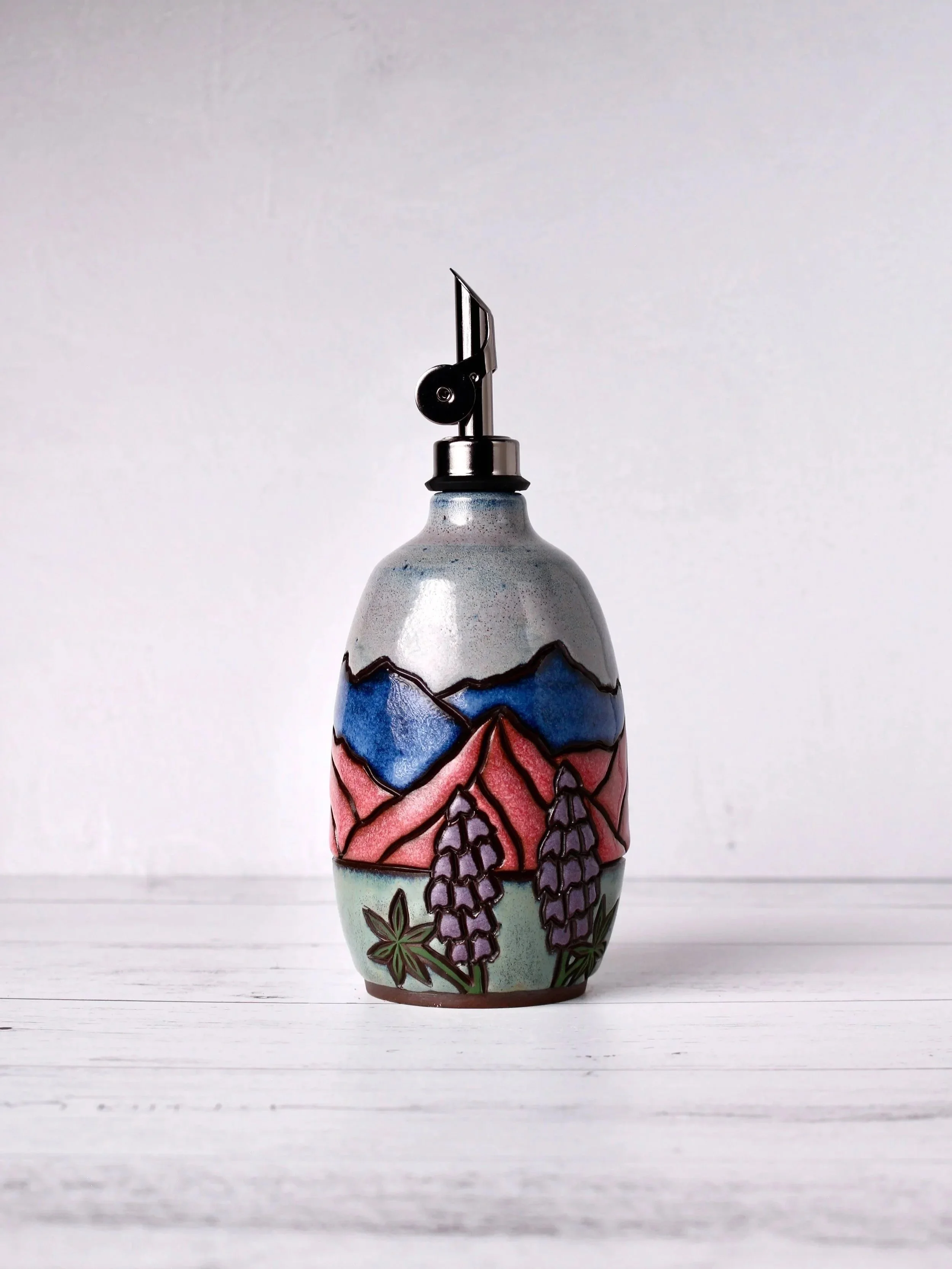 Decorative ceramic oil pourer with floral mountain design and purple wisteria flowers, placed on a white wooden surface.