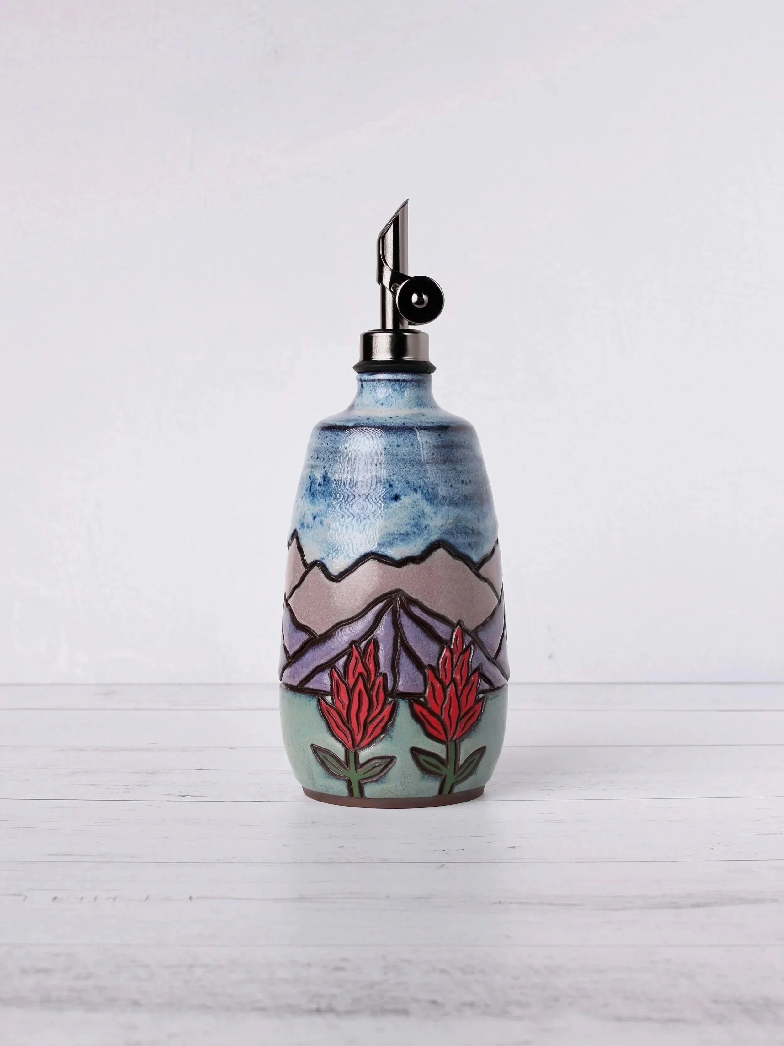 A ceramic vase with mountain, tree, and floral designs painted on it, featuring a metal pour top, set against a white background.
