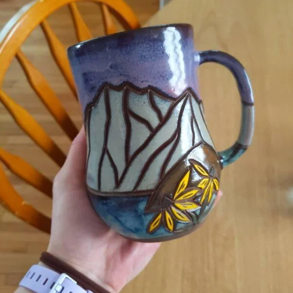 Colorful ceramic mug decorated with mountain and leaf designs, being held in a person's hand over a wooden table.