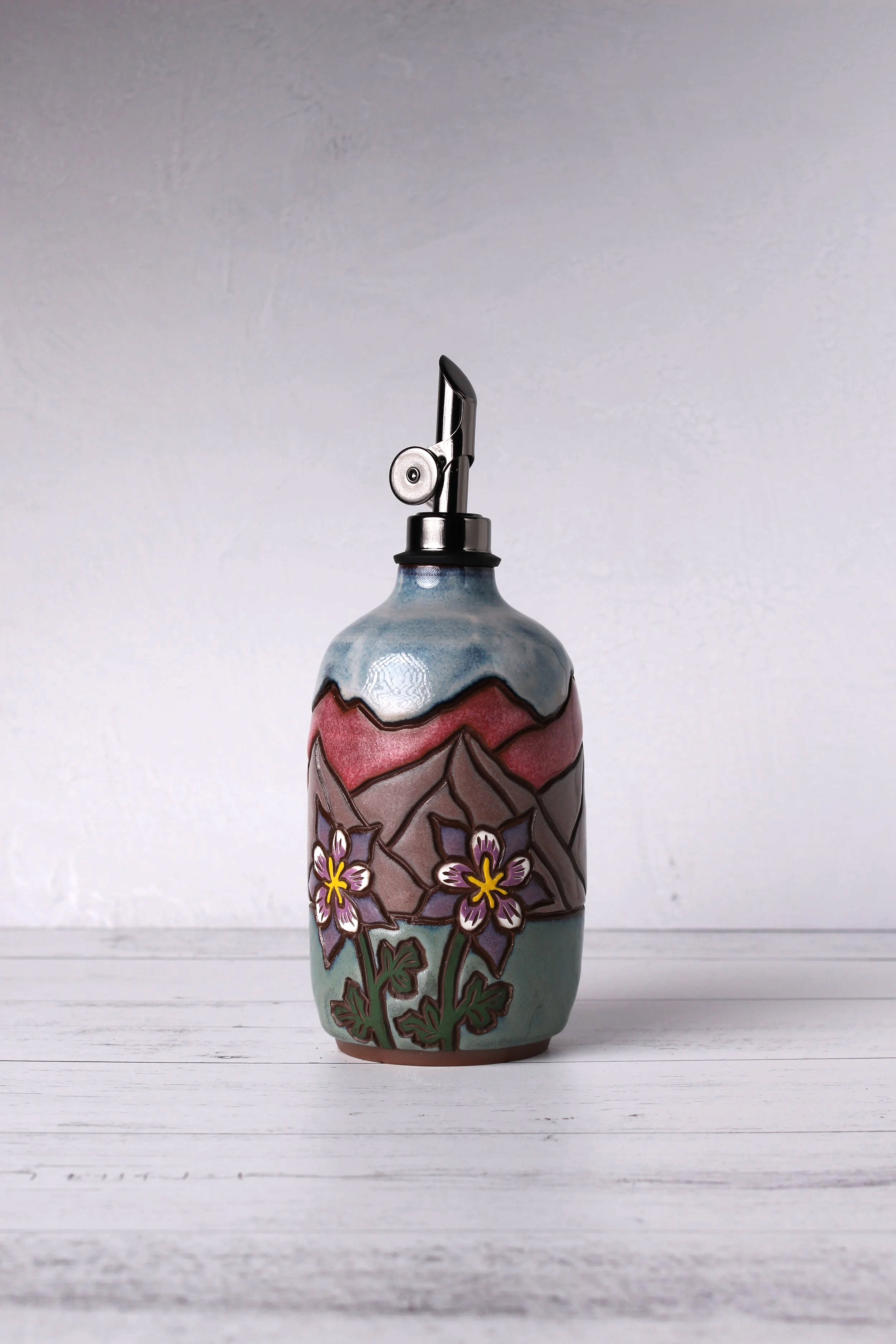 A decorative ceramic oil bottle with floral and plant patterns, topped with a metal spout, set against a plain, light-colored background.