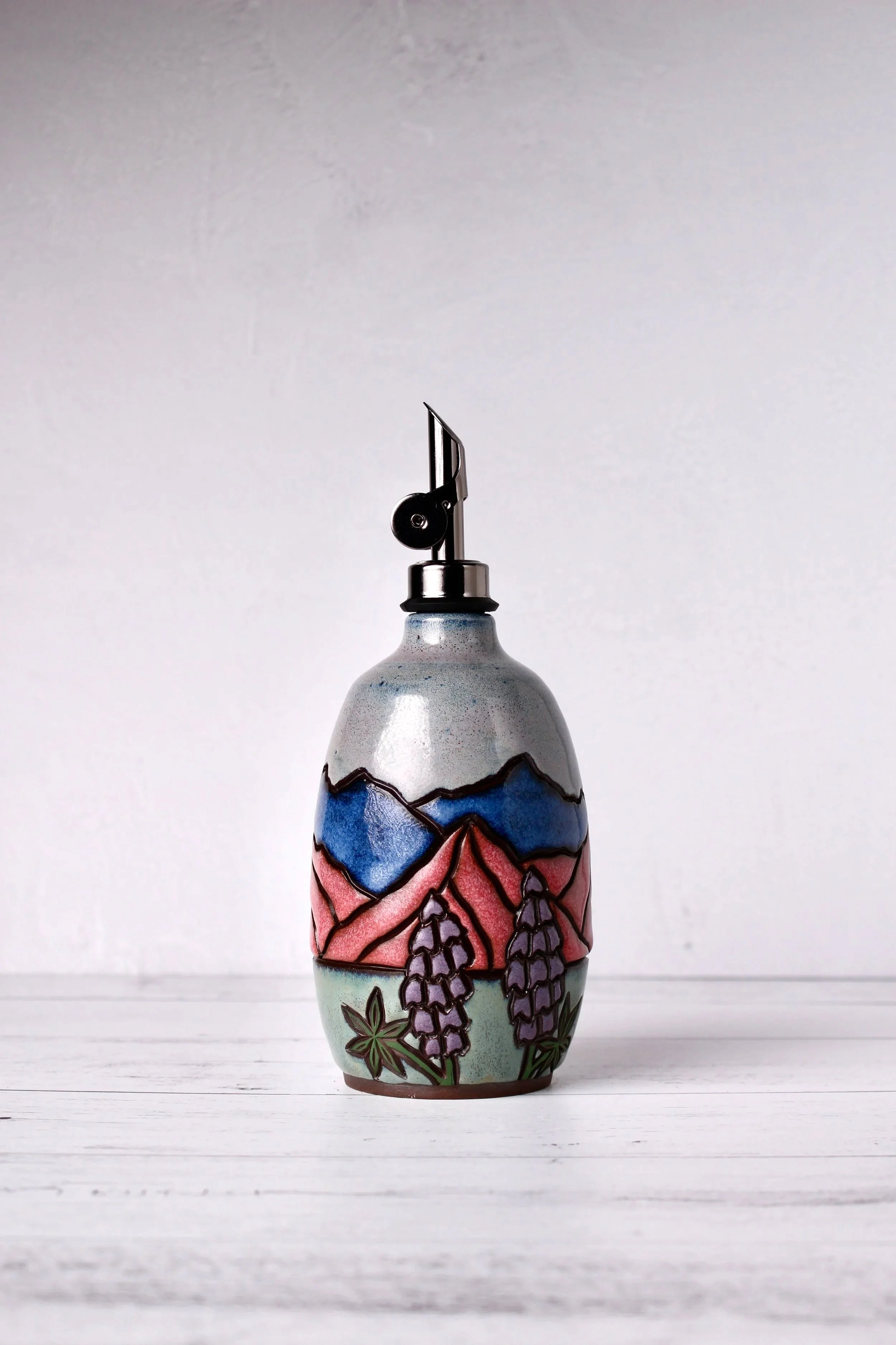 Colorful ceramic oil vessel with a painted landscape scene including mountains, trees, and flowering plants, placed on a white wooden surface.