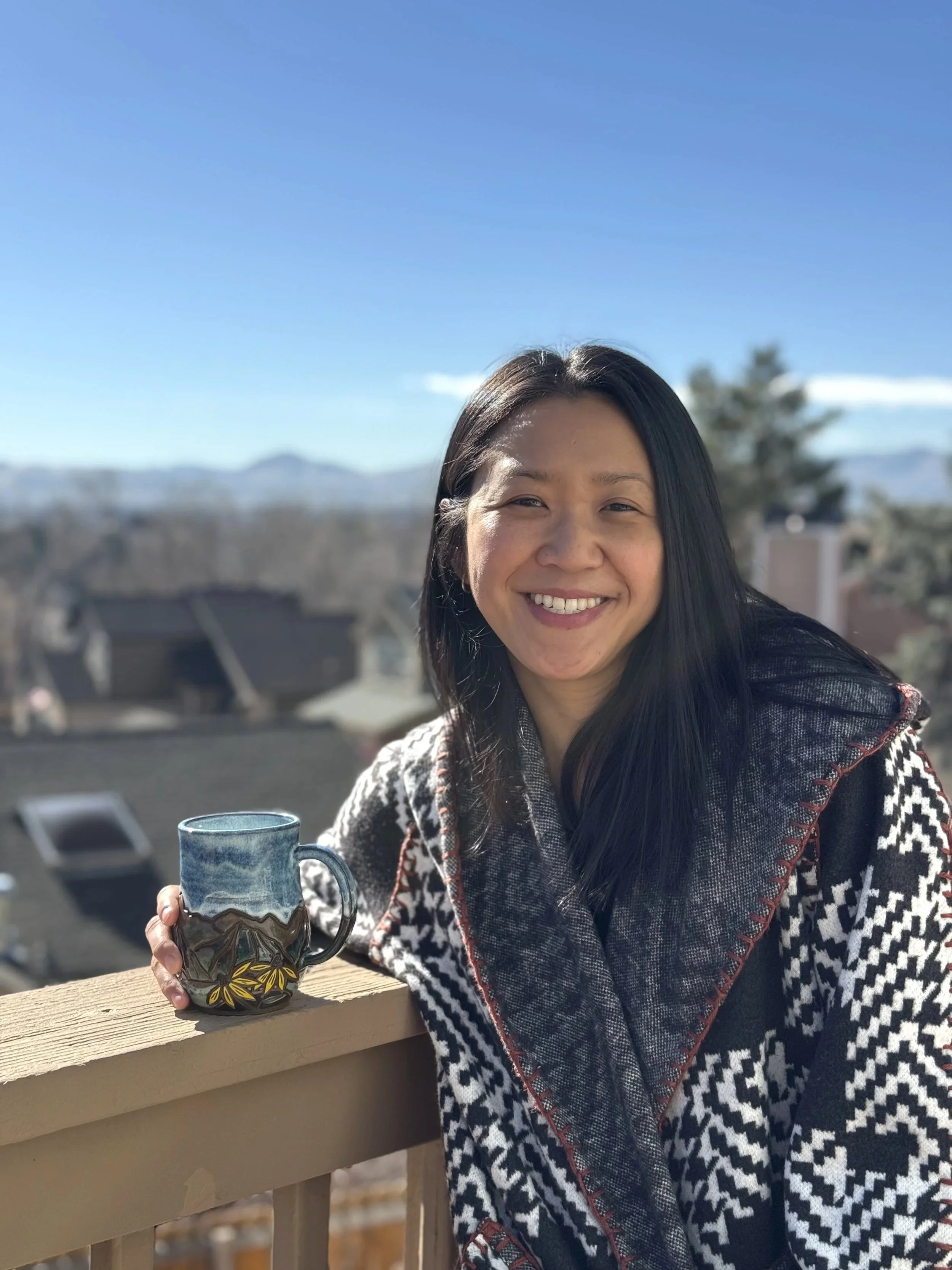 A smiling woman with long black hair holding a ceramic mug with a colorful design, standing outdoors on a bright, sunny day with mountains and a house in the background.
