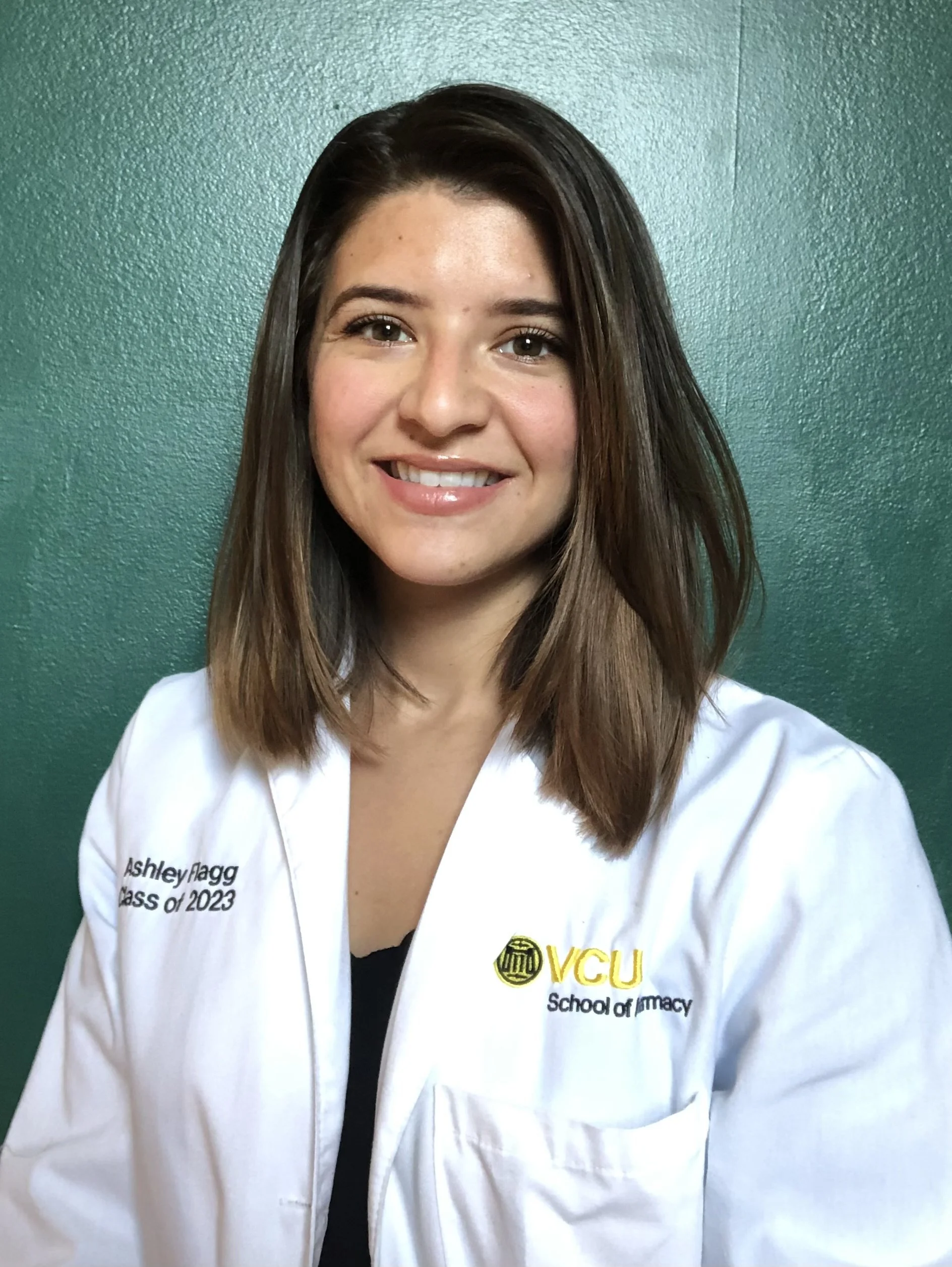 Young woman with long brown hair, smiling, wearing a VCU School of Pharmacy white lab coat standing against a green wall.