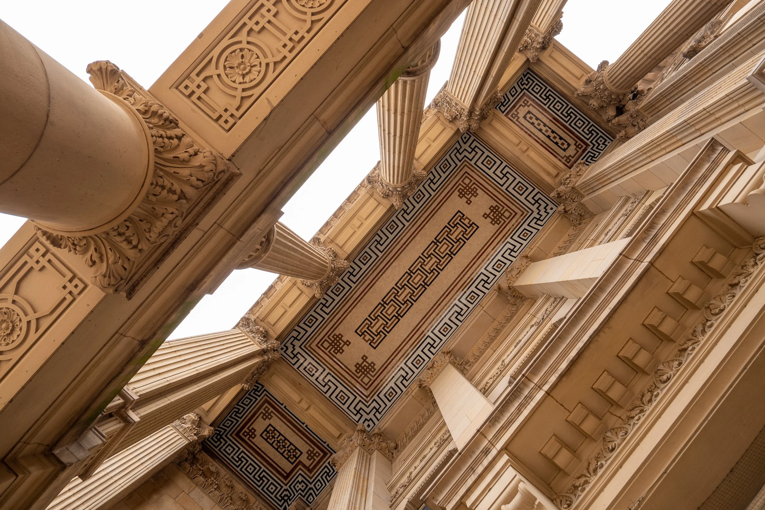 Looking up at the ornate underside of a classical building with decorative columns, intricate carvings, and geometric patterns on the ceiling, viewed from below.