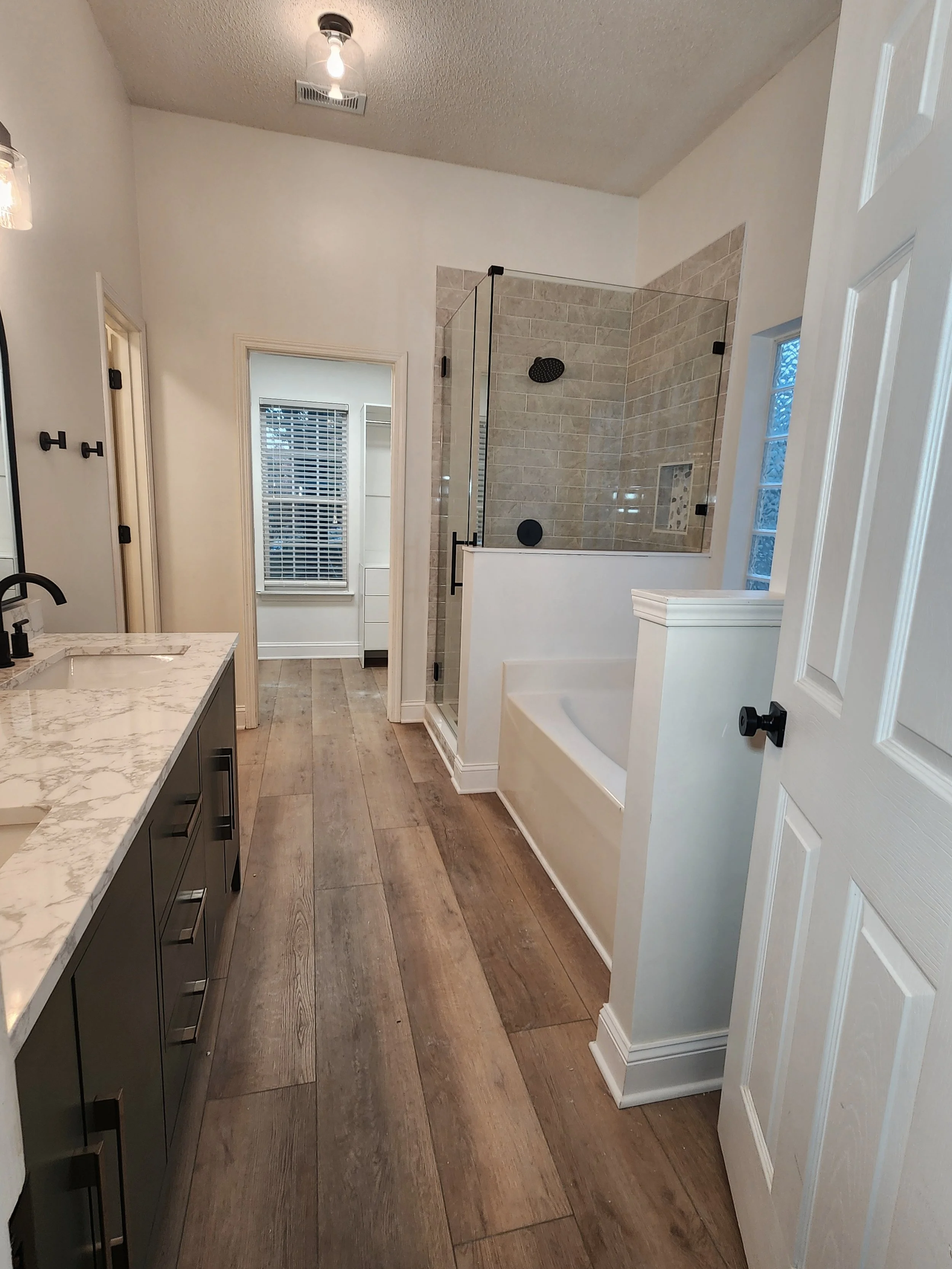 A modern bathroom with a wooden floor, a dark vanity with drawers, a white marble countertop, a sink with a black faucet, a glass-enclosed walk-in shower with beige tiled walls, a built-in niche in the shower, and a window with white blinds.
