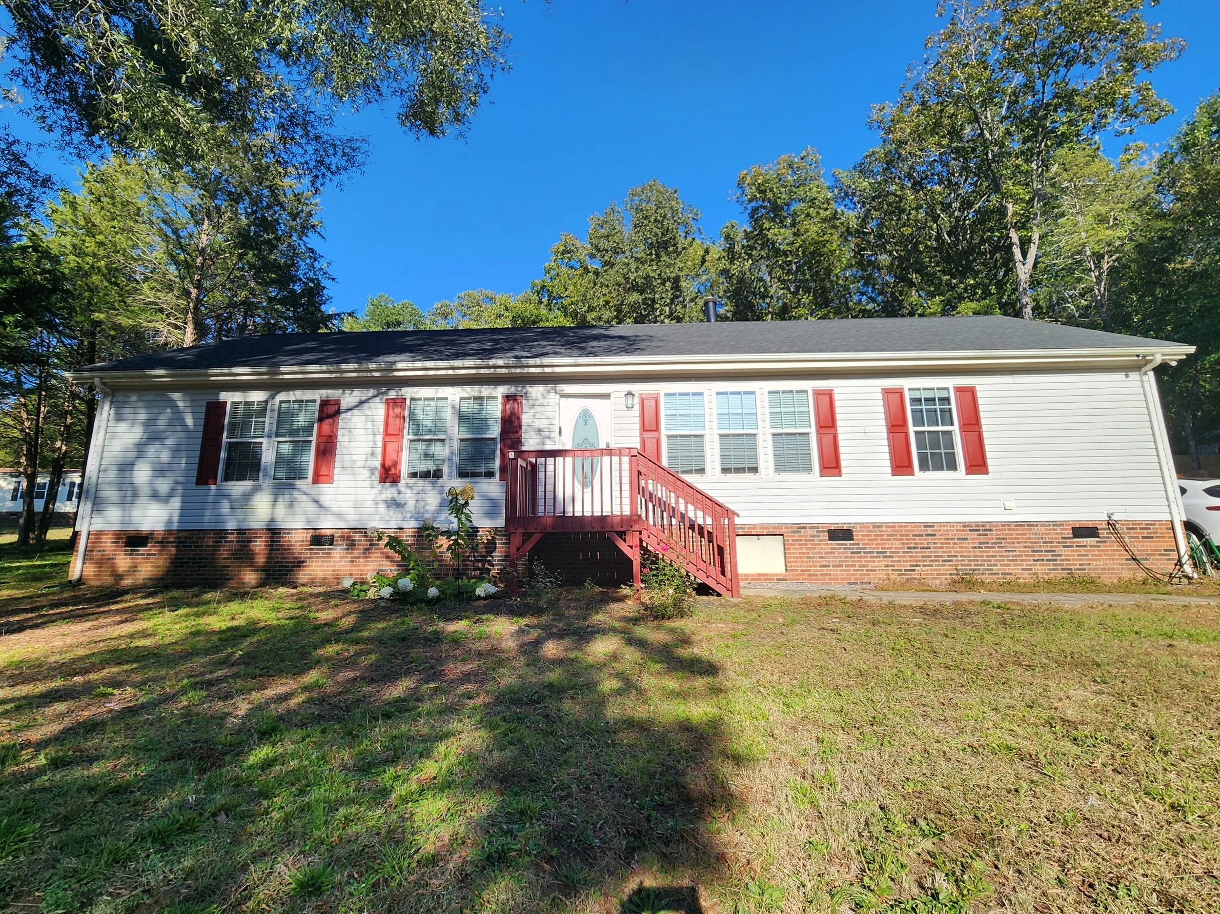 A single-story house with white siding and red shutters, a black roof, and a red staircase leading to the front door, surrounded by green trees and a grassy yard.