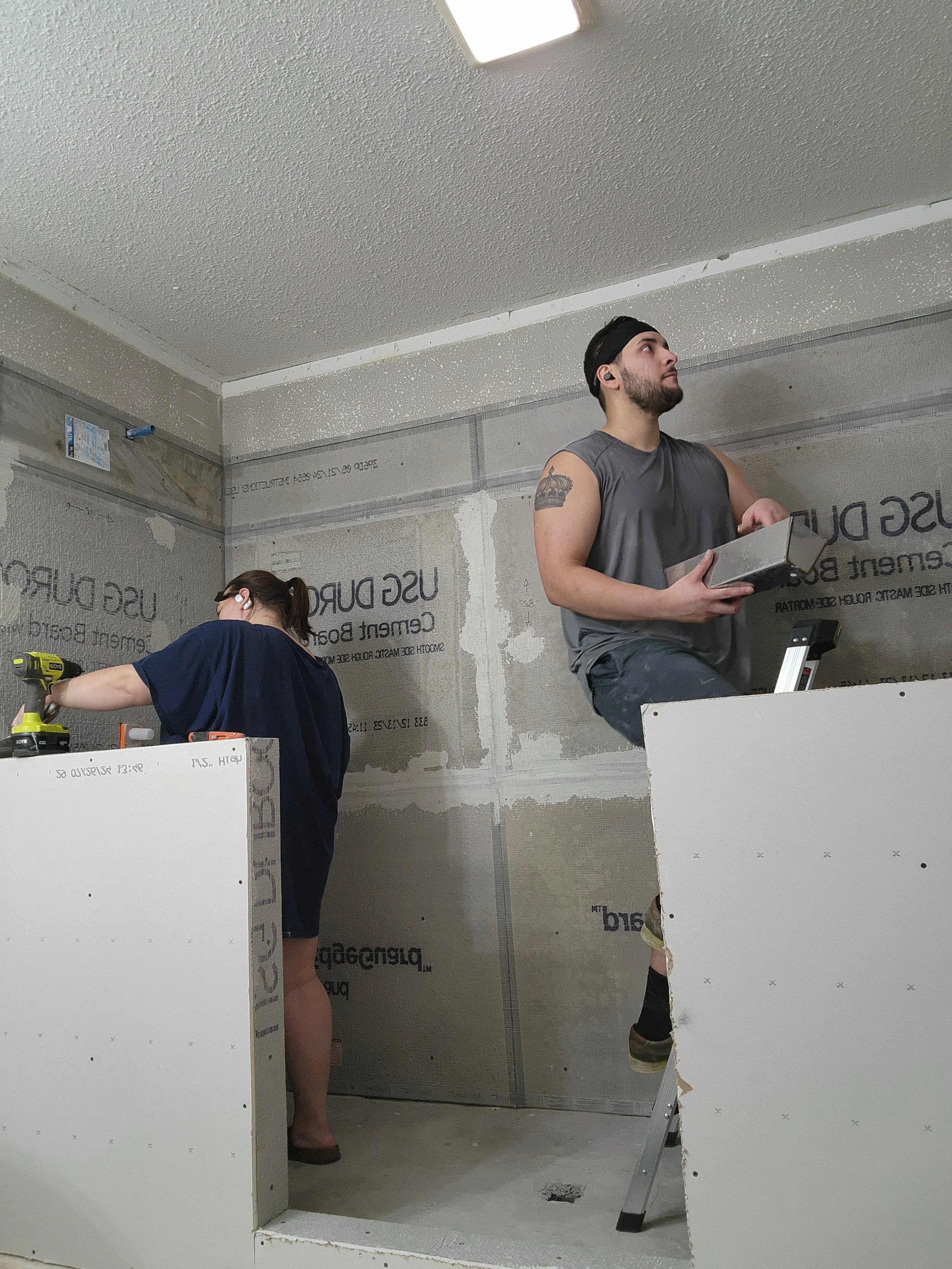 Two people working in an unfinished room with drywall. One person, a man, is sitting on a stepladder holding a box, while the other person, a woman, is standing and using a power drill on the drywall.