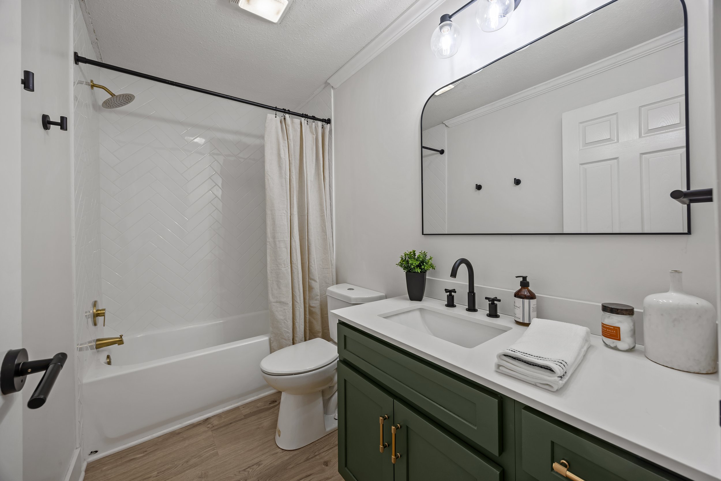 A modern bathroom with a white bathtub and shower, a white toilet, a green vanity with gold handles, a large mirror, and decorative items like towels, a plant, and soap bottles. The shower has a black and gold showerhead and fixtures, with a black shower curtain rod and hooks. The flooring is wood-like tile.