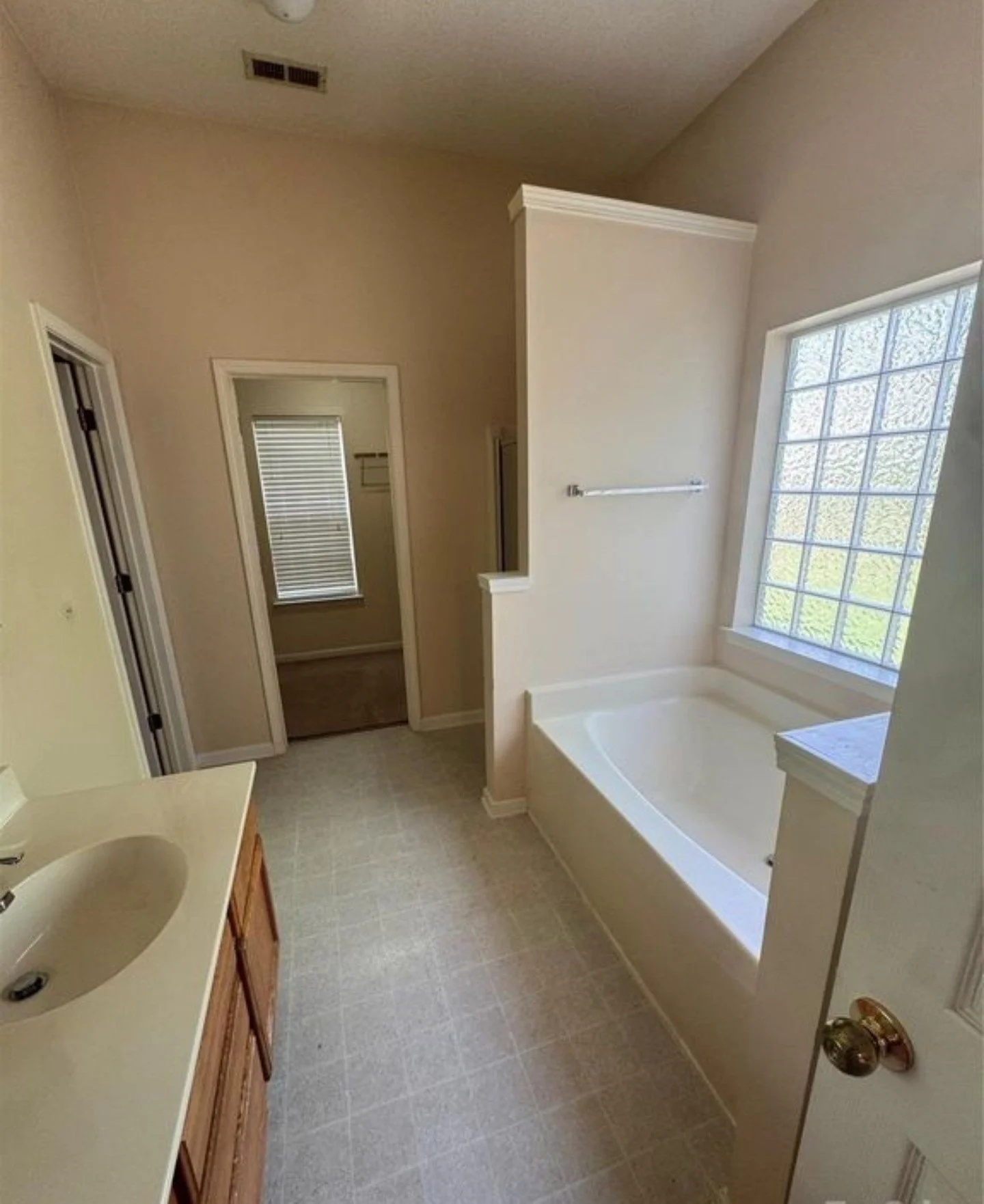 A bathroom with a large window, bathtub, vanity with two sinks, and a door leading to another room with window blinds.