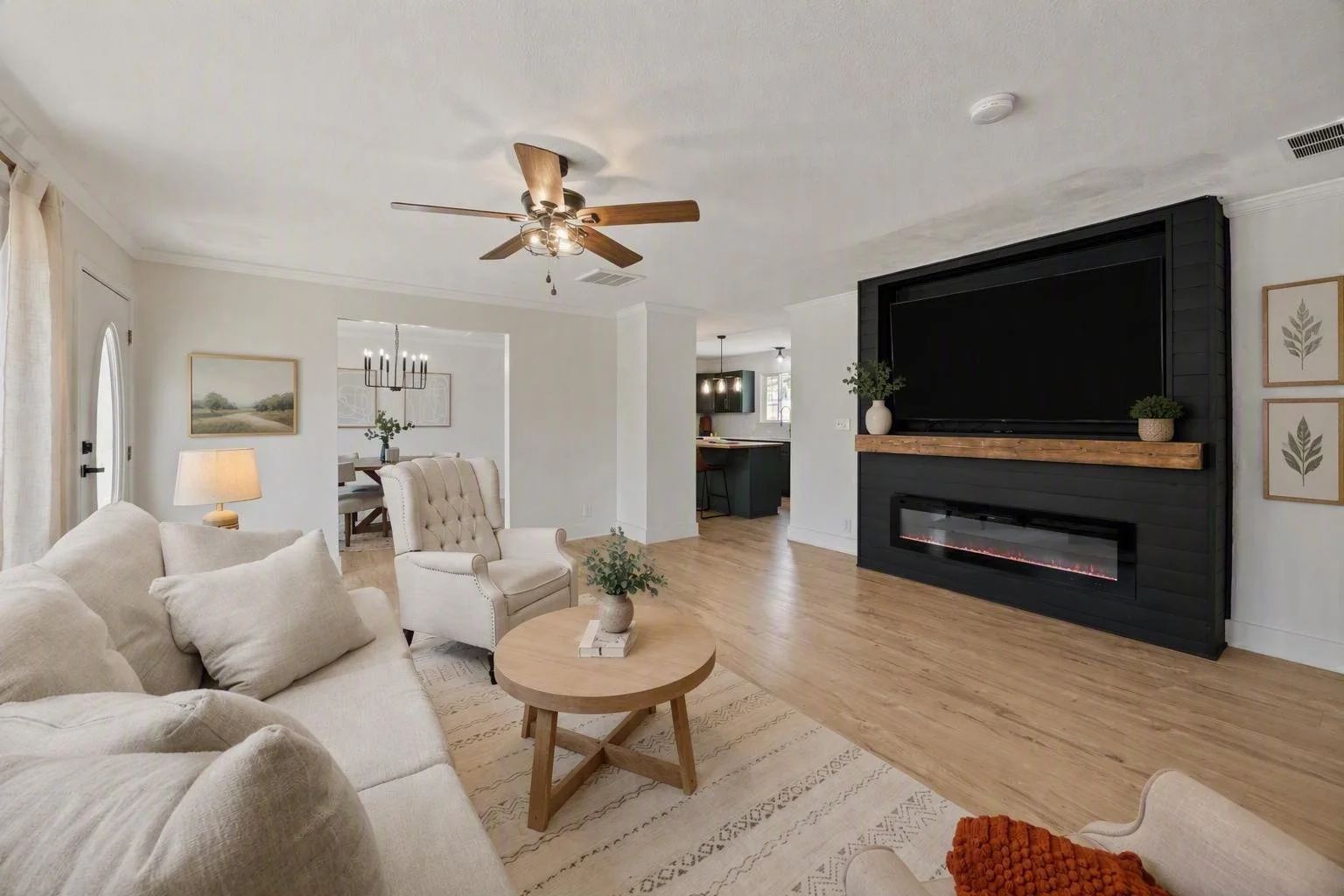 Living room with white sofa, armchair, round wooden coffee table, black television above electric fireplace, wooden mantle, ceiling fan, artwork on walls, and hardwood floors.