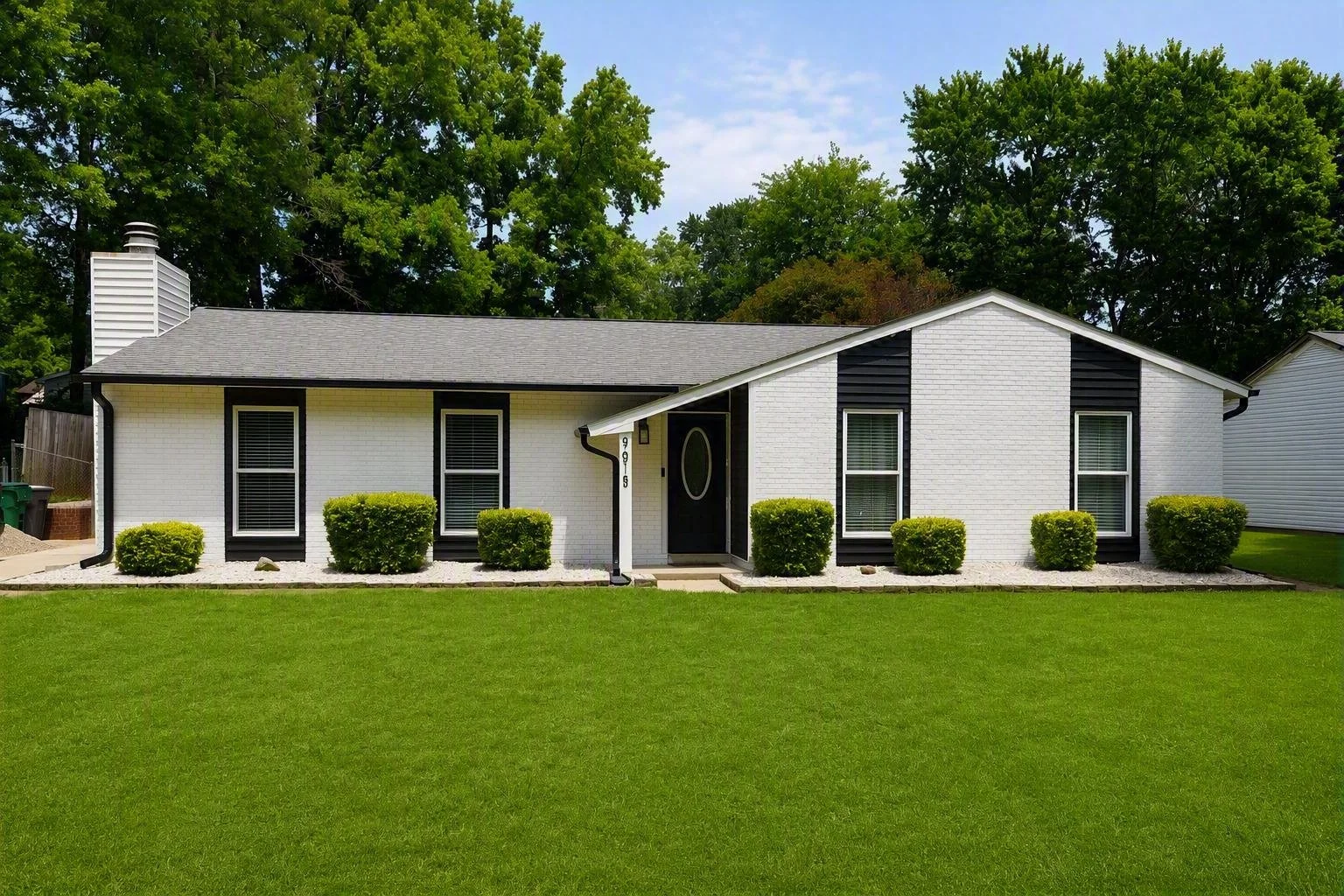 Front view of a white brick house with black shutters, black front door, and neatly trimmed bushes in the yard, with tall trees in the background.