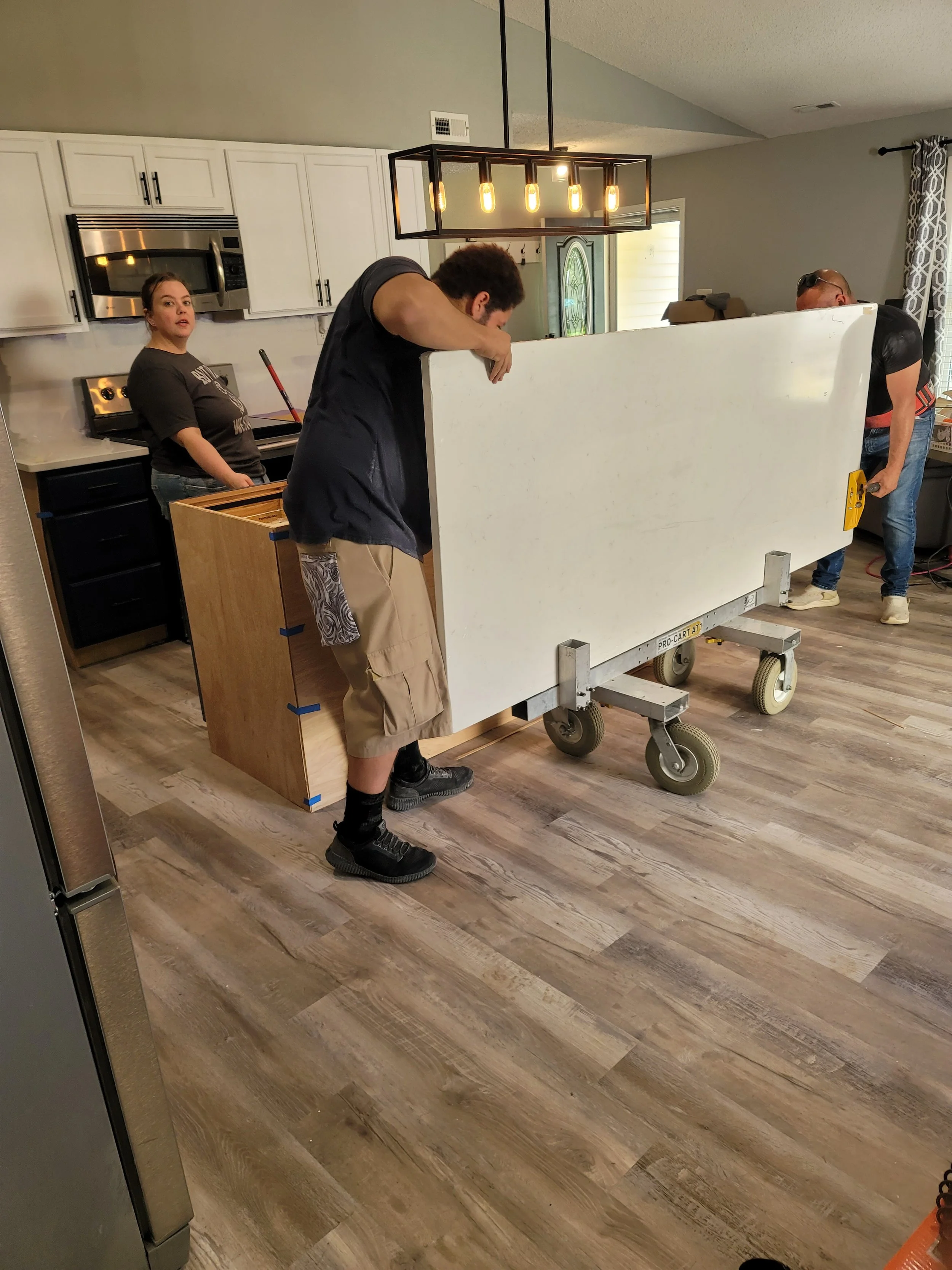 People working on a kitchen island, with one person inspecting the side of the island and another person looking at the front. A woman is standing near the stove in the background.