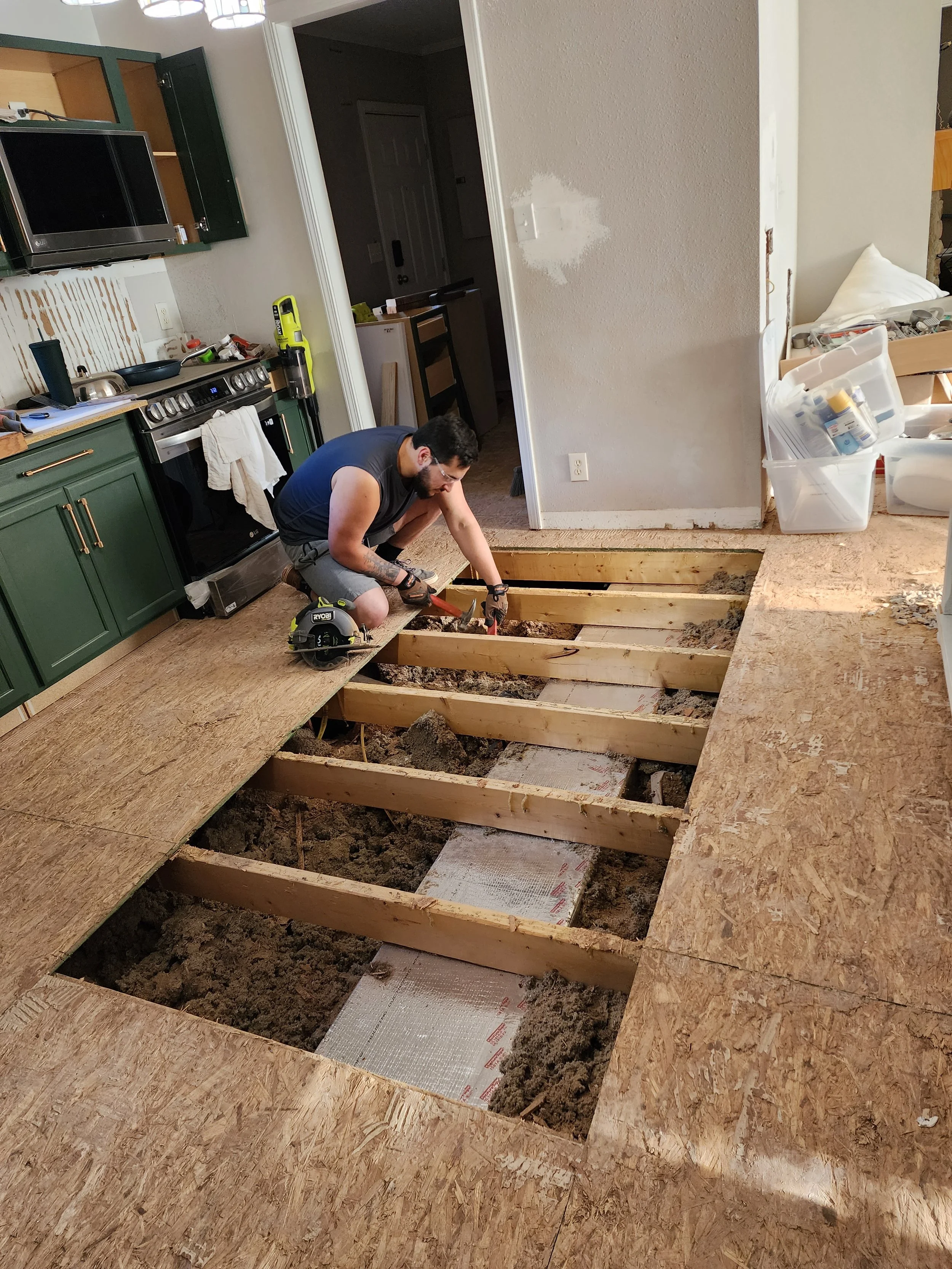 A man is kneeling on a wooden floor frame in a kitchen, working on installing flooring. The room has been partially torn up, with exposed joists and insulation, and construction materials are visible.
