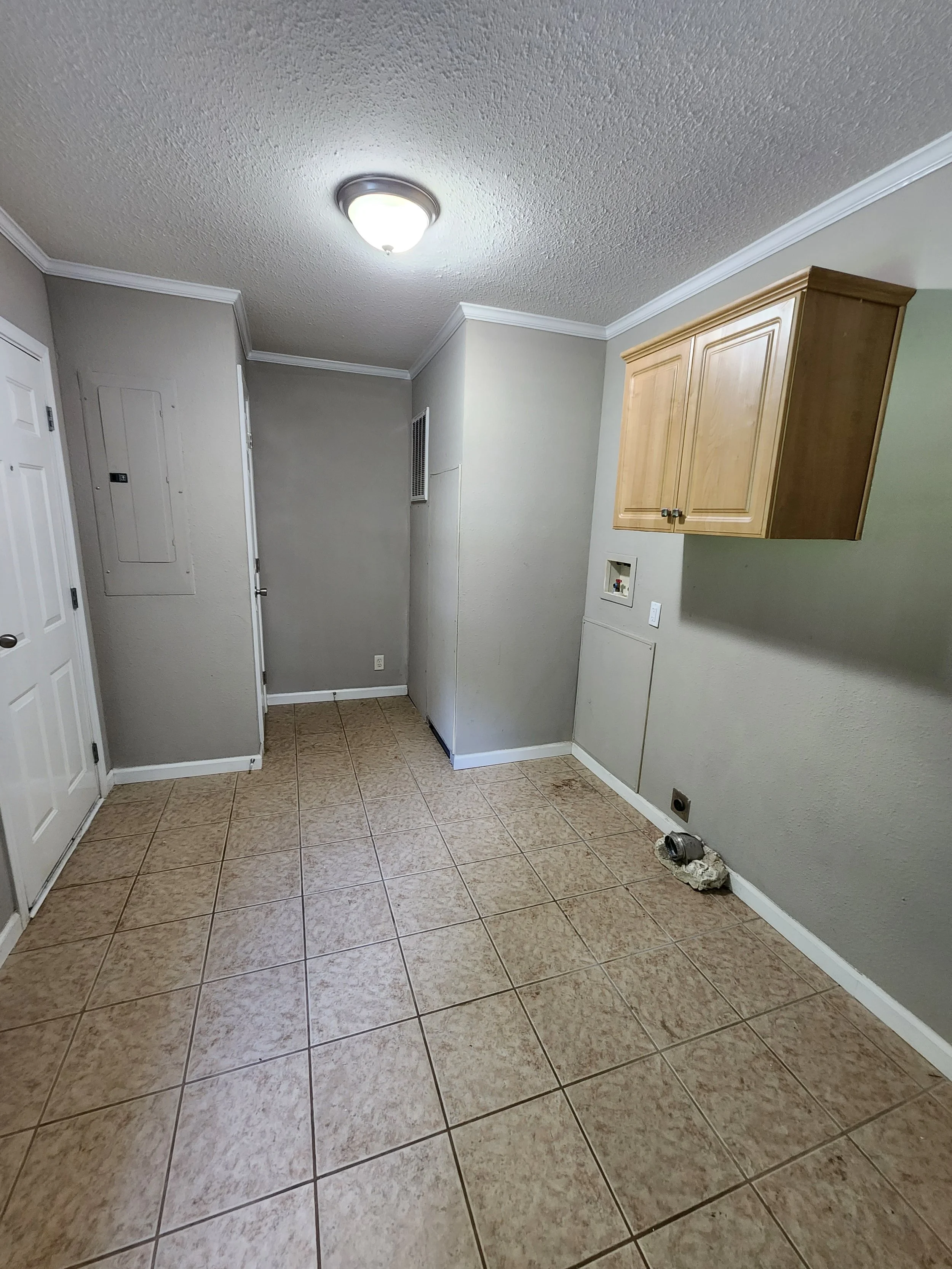 Empty room with beige tiled floor, beige walls, and wooden cabinets. Part of the wall has a small opening with exposed pipes and debris on the floor.