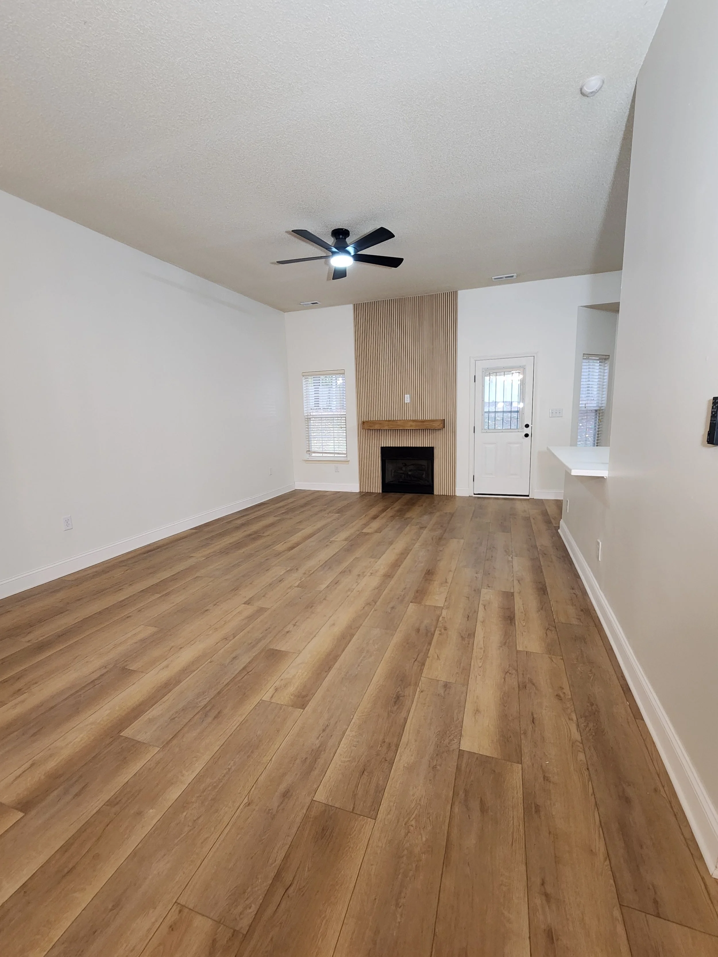 Living room with wooden floor, white walls, a ceiling fan, and a fireplace on a wall with vertical wooden paneling, two windows, and a door.