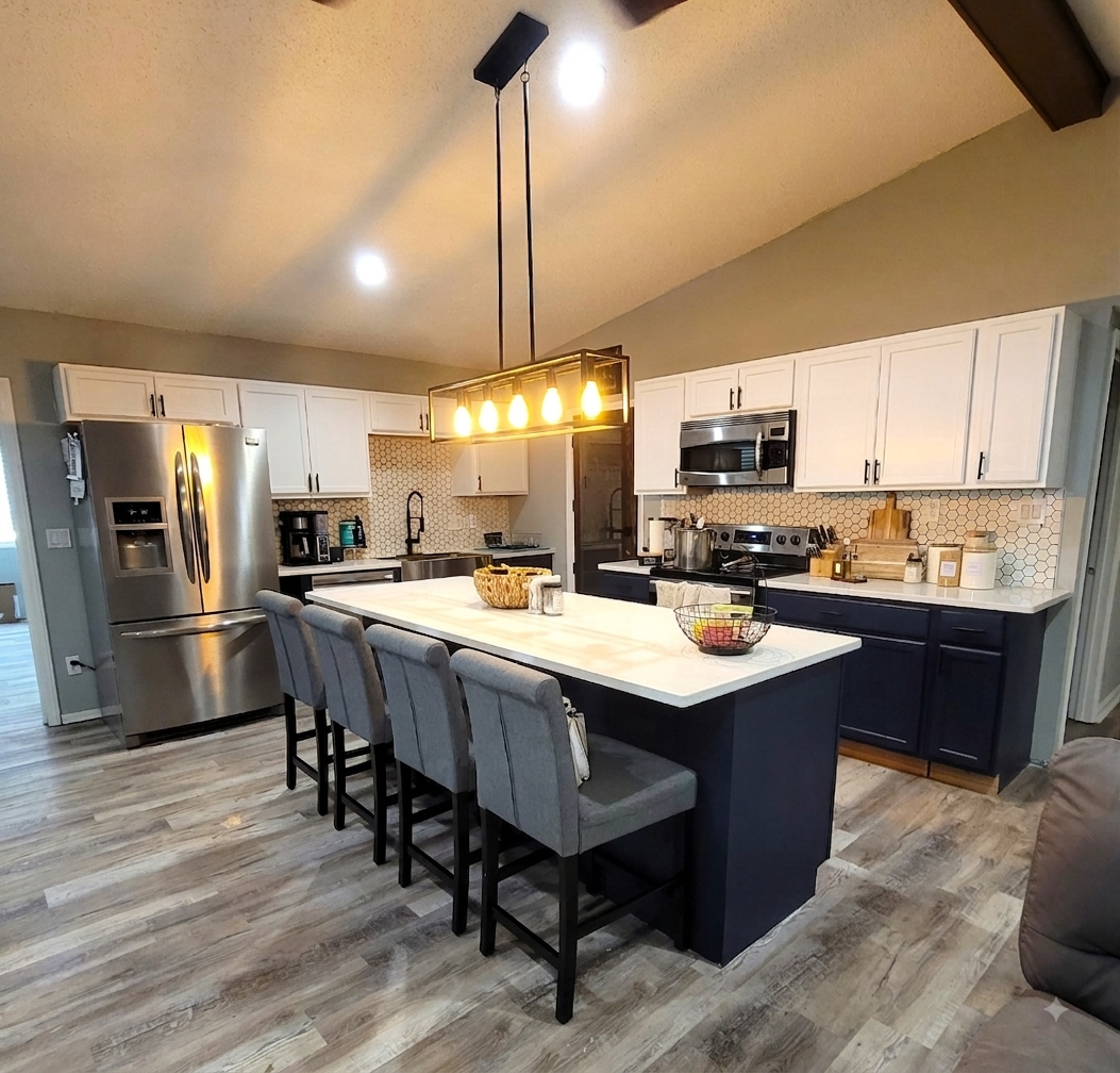 Modern kitchen with white upper cabinets, navy lower cabinets, and stainless steel appliances. Kitchen island with a white countertop, gray bar stools, and a black and gold pendant light.