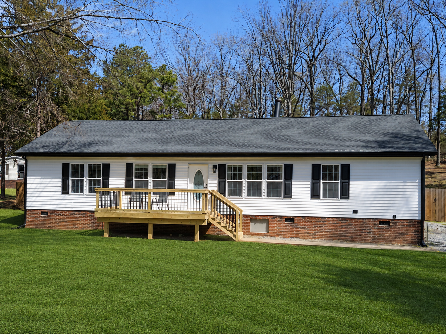 A house with white siding, black shutters, a gray roof, and a brick foundation. It has a small wooden deck with stairs and a grassy yard.