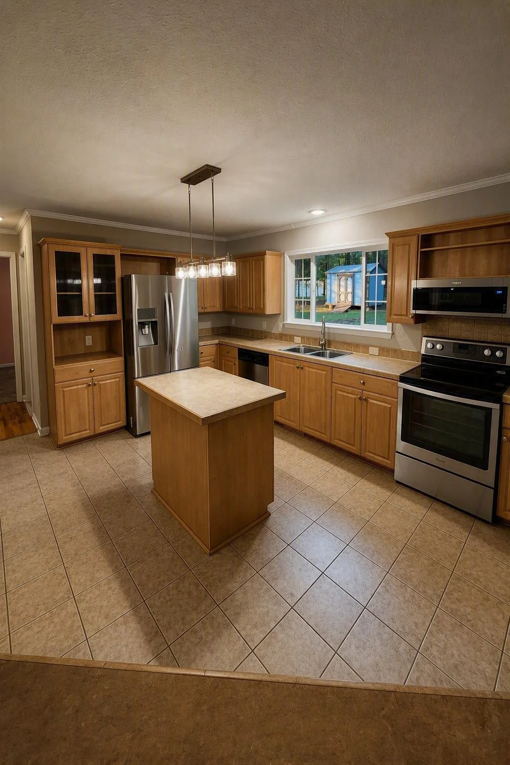 Kitchen with wooden cabinets, stainless steel refrigerator, stove, microwave, and a small island in the center. A window shows a backyard with a shed.