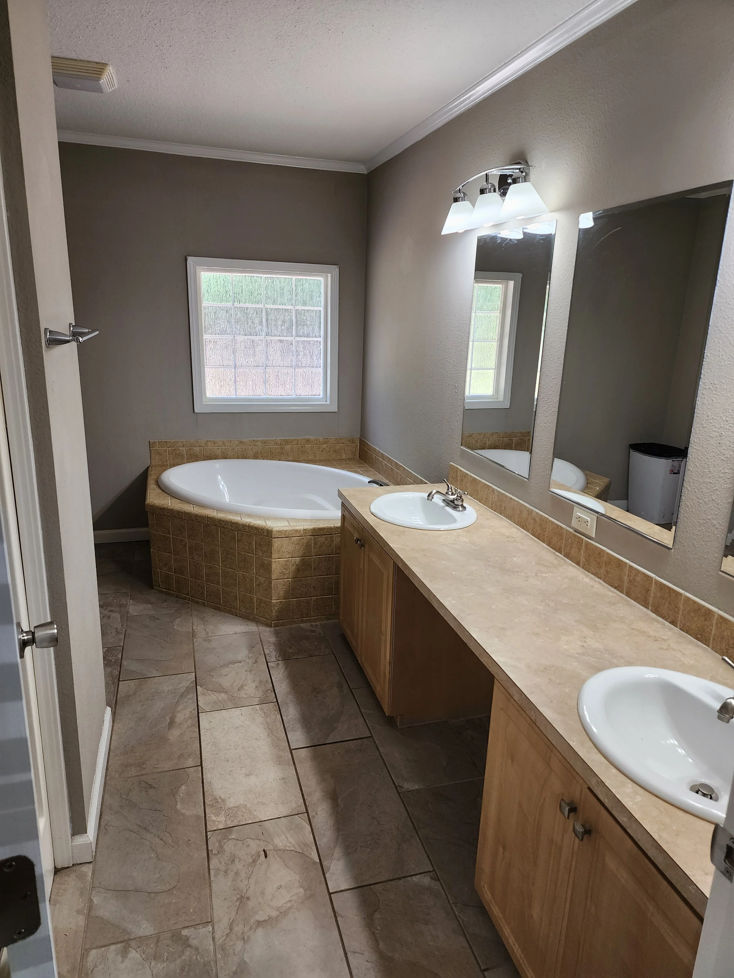 A bathroom with a large window, a bathtub with a tile surround, a double sink vanity with a marble countertop, a mirror, and overhead light fixtures.