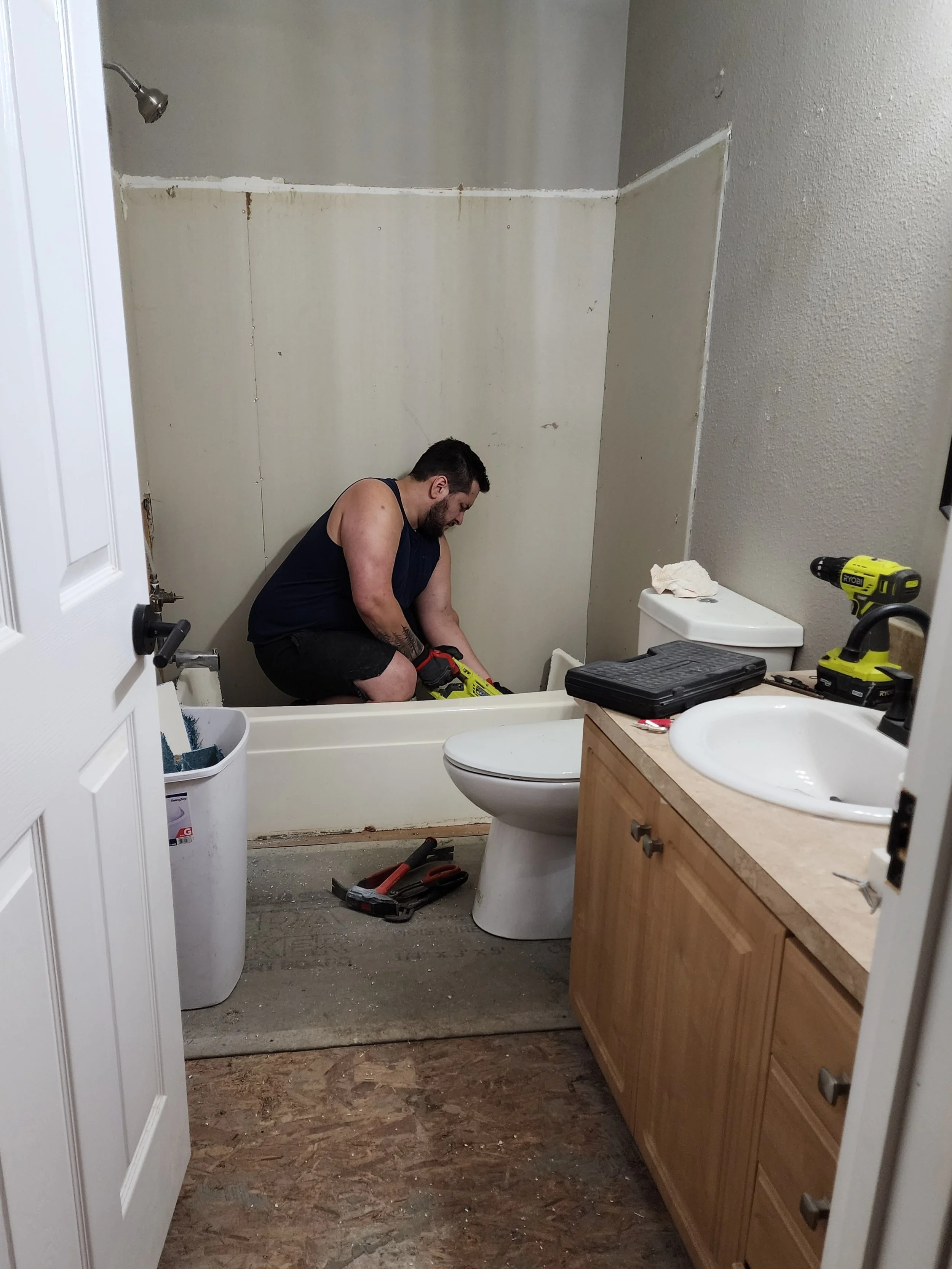 A man is working on a bathroom renovation in a room with a bathtub and a vanity. The walls are partially finished, with some drywall and paint. Tools and construction materials are scattered around, and the floor is exposed plywood.