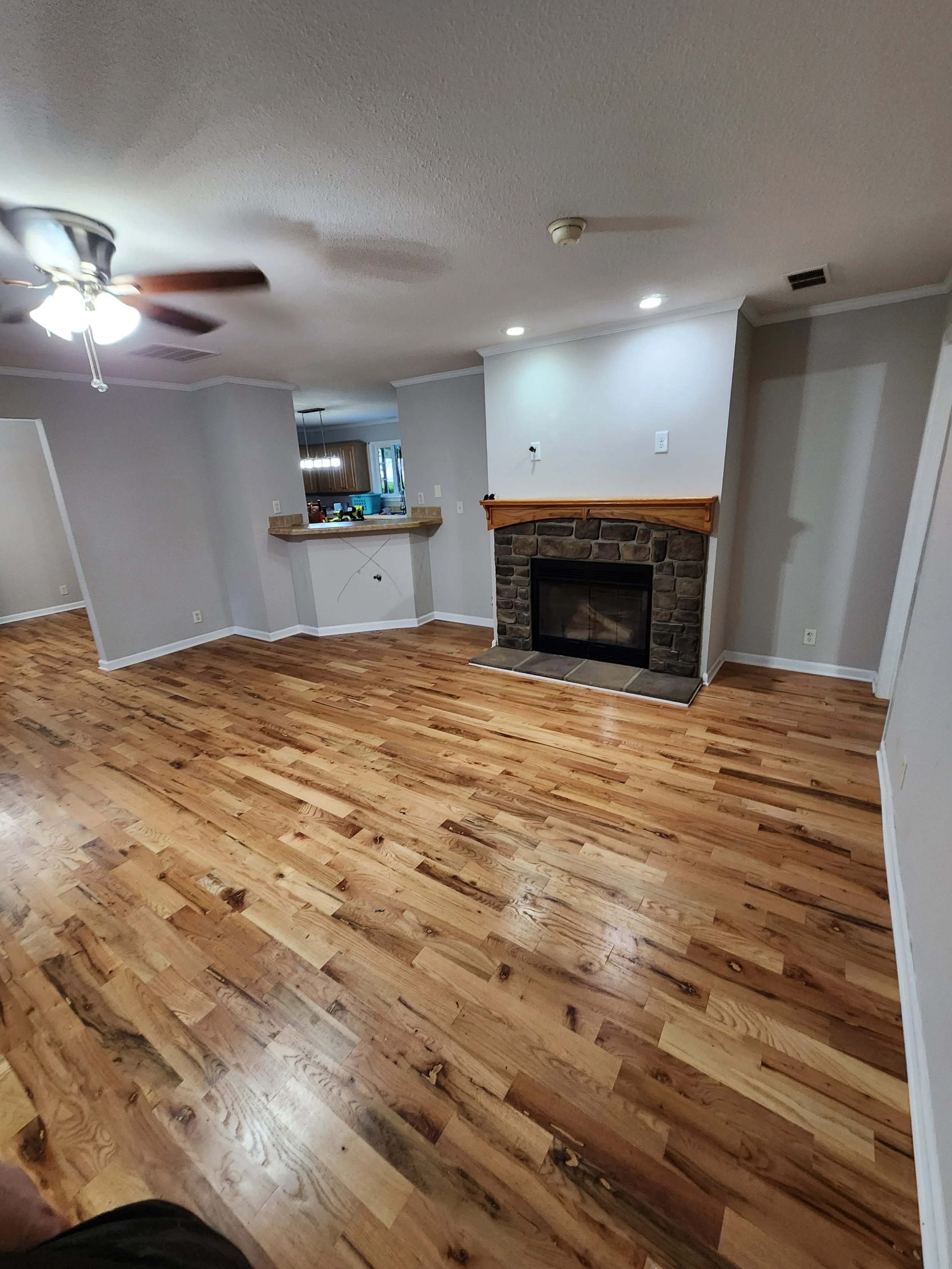 Living room with wooden floor, white walls, ceiling fan, fireplace with stone and wooden mantel, and kitchen visible in background.