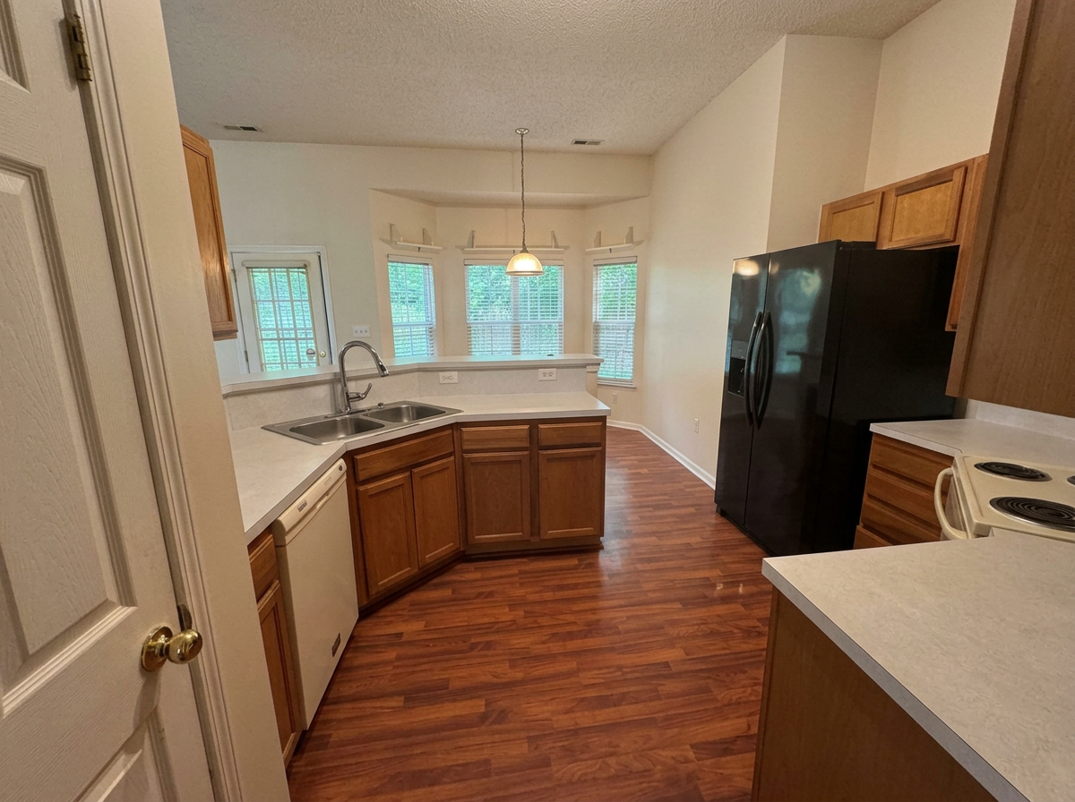Kitchen with wooden cabinets, black refrigerator, white stove, and a double sink, with a dining area with bay windows and a hanging light.