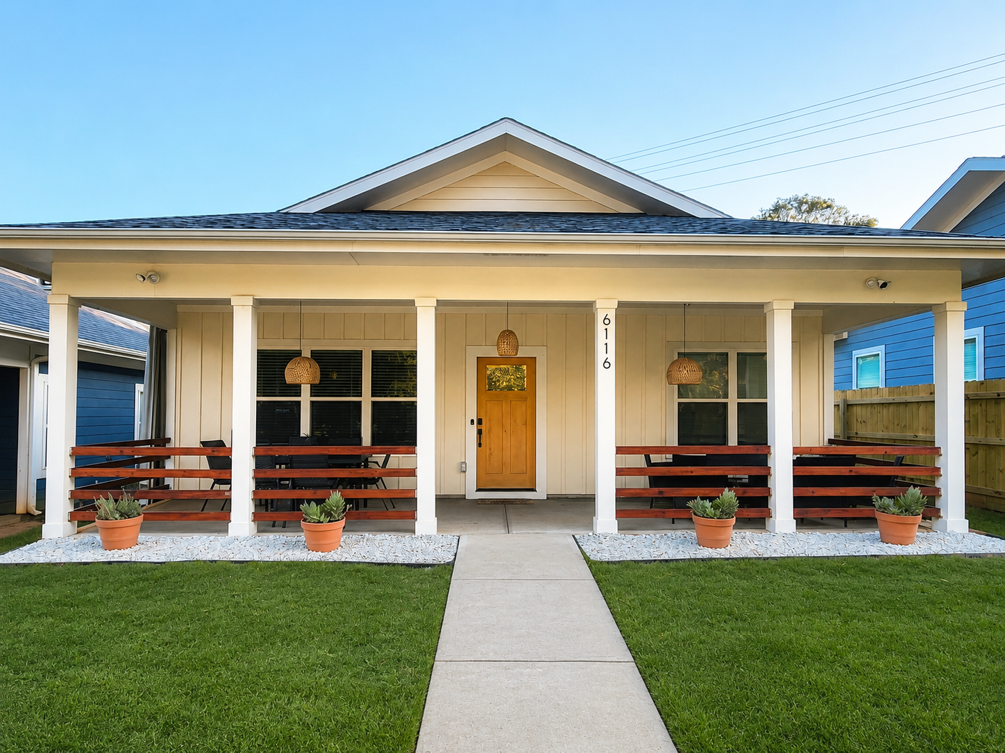 Front view of a modern house with a white exterior, wooden front door, and a front porch with four seats. Potted plants are placed along the front, and the house number 6116 is displayed on a white column. The lawn is green, and a concrete walkway leads to the front door.
