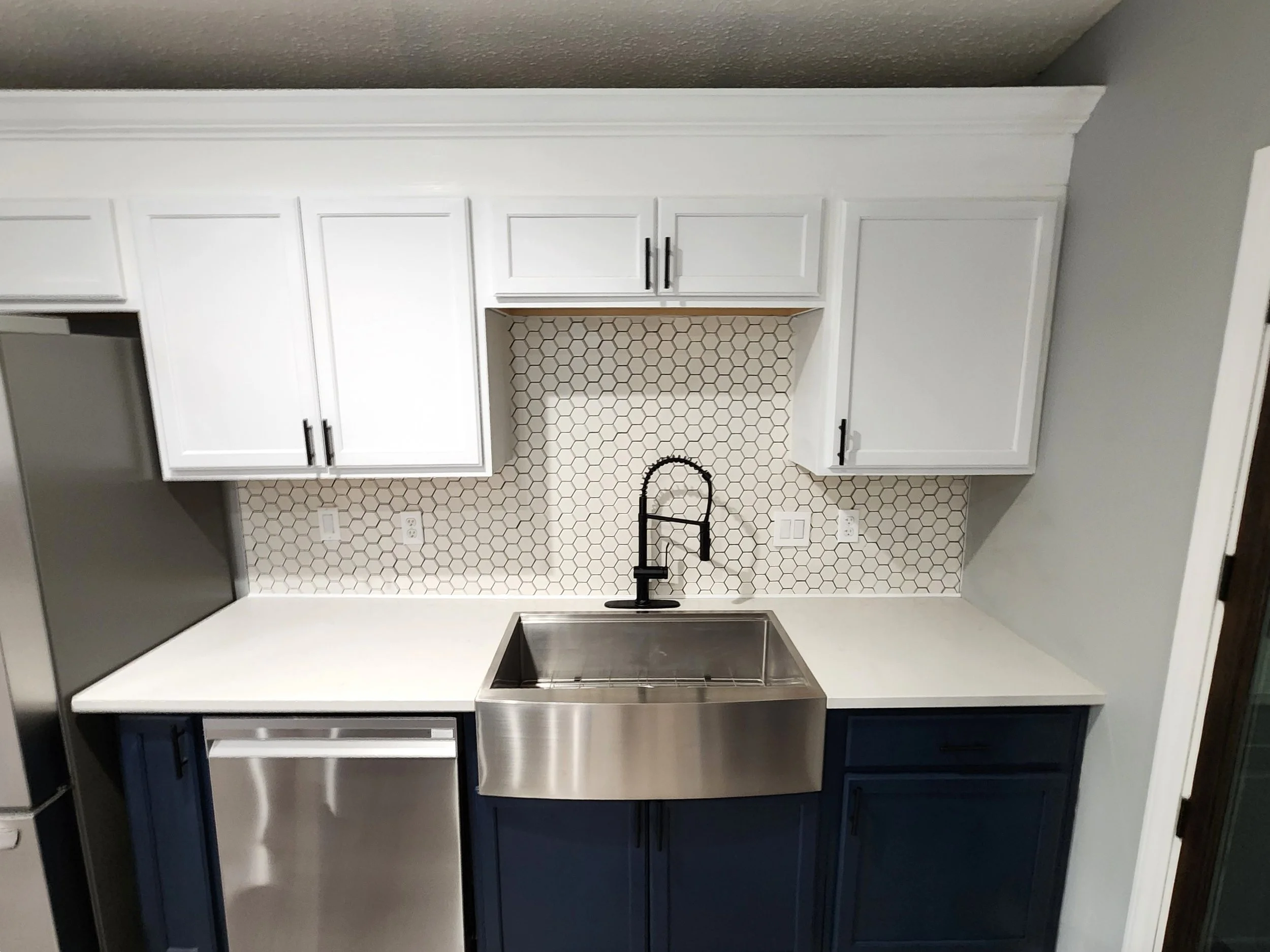 Modern kitchen with white upper cabinets, dark lower cabinets, a stainless steel sink, black faucet, and a hexagon tile backsplash.