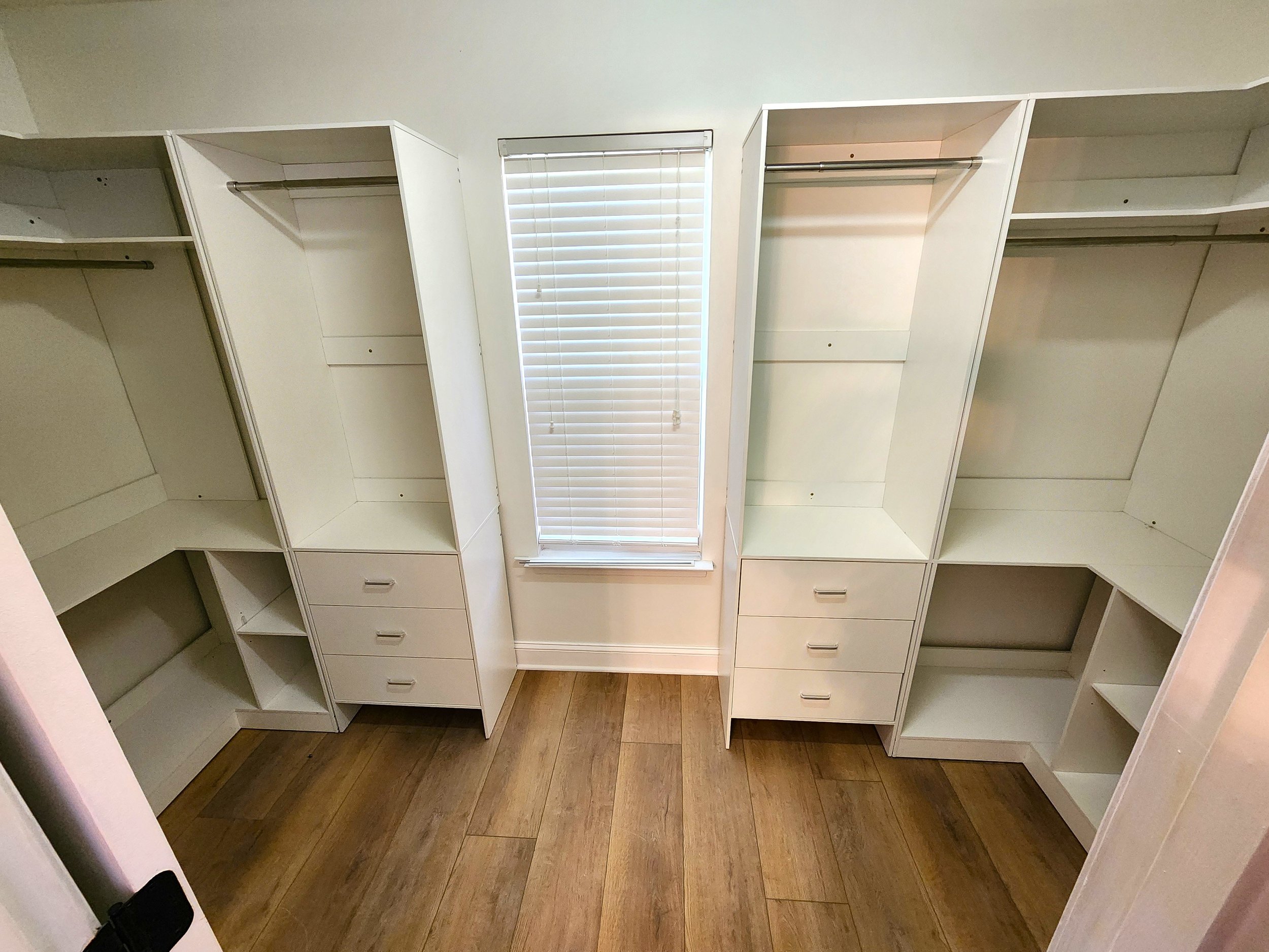 Empty white built-in closet with multiple shelves and drawers, a window with blinds, and wood flooring in a room.