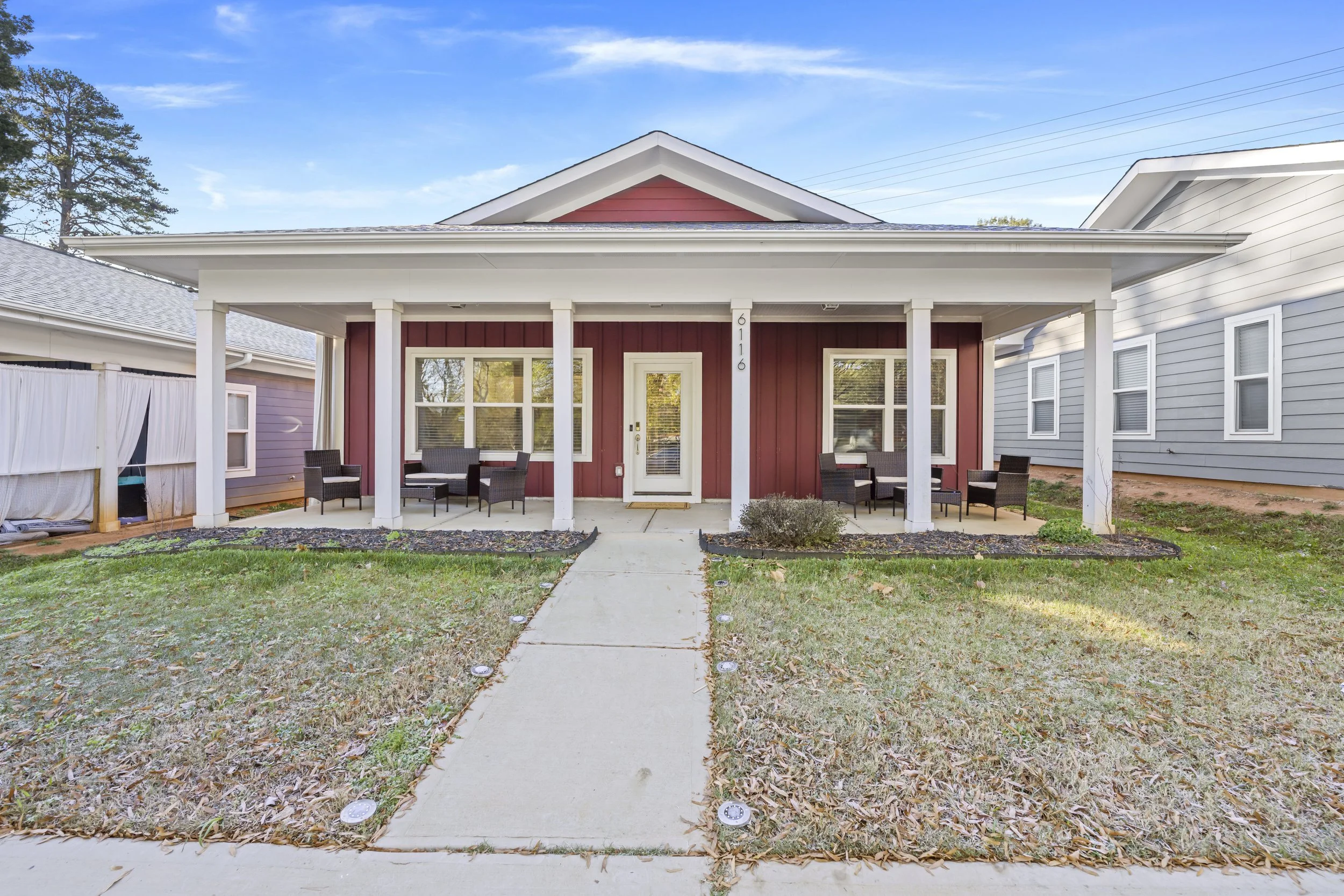 Front view of a small house with a porch, red siding, white trim, and outdoor seating, surrounded by grass and neighboring houses under a blue sky.