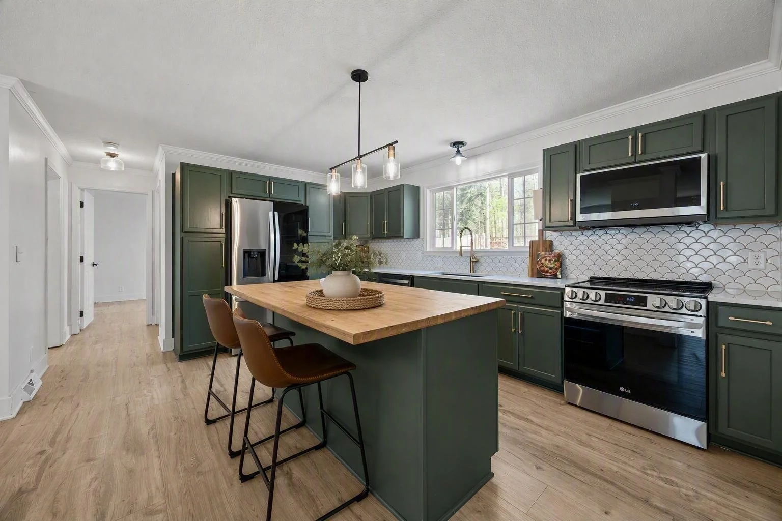 Modern kitchen with dark green cabinets, stainless steel appliances, a wooden island with brown chairs, a black pendant light, and a large window above the sink.