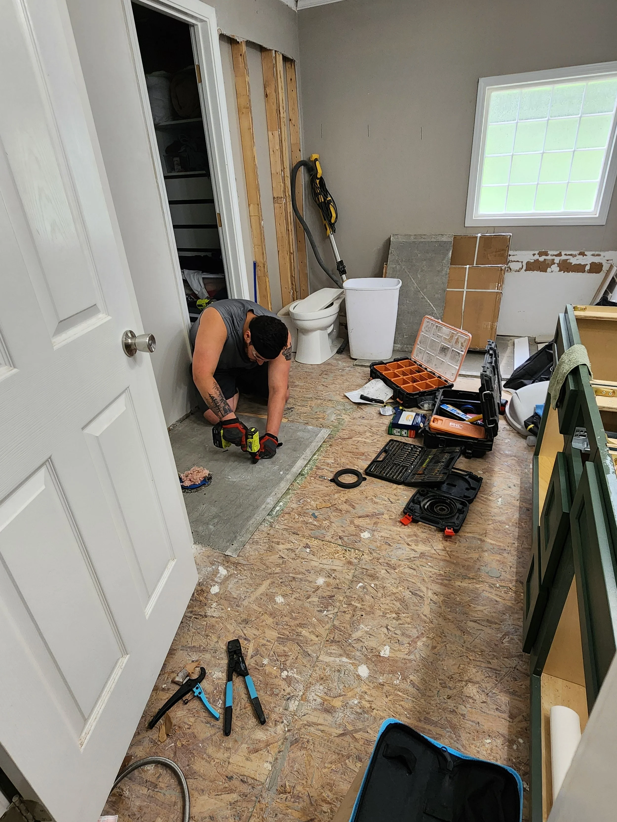 Person kneeling on the floor of a bathroom renovation, working with tools surrounded by construction materials, while a window, toilet, and partially removed wall are visible.