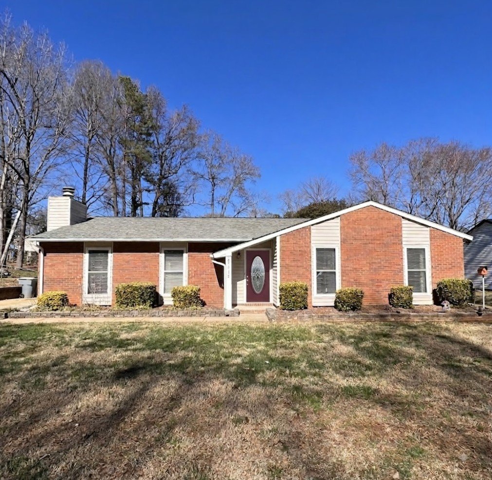 Single-story brick house with a gray shingle roof, white trim, and a red front door with a decorative oval window. There are grass and bushes in the front yard, and leafless trees in the background under a clear blue sky.