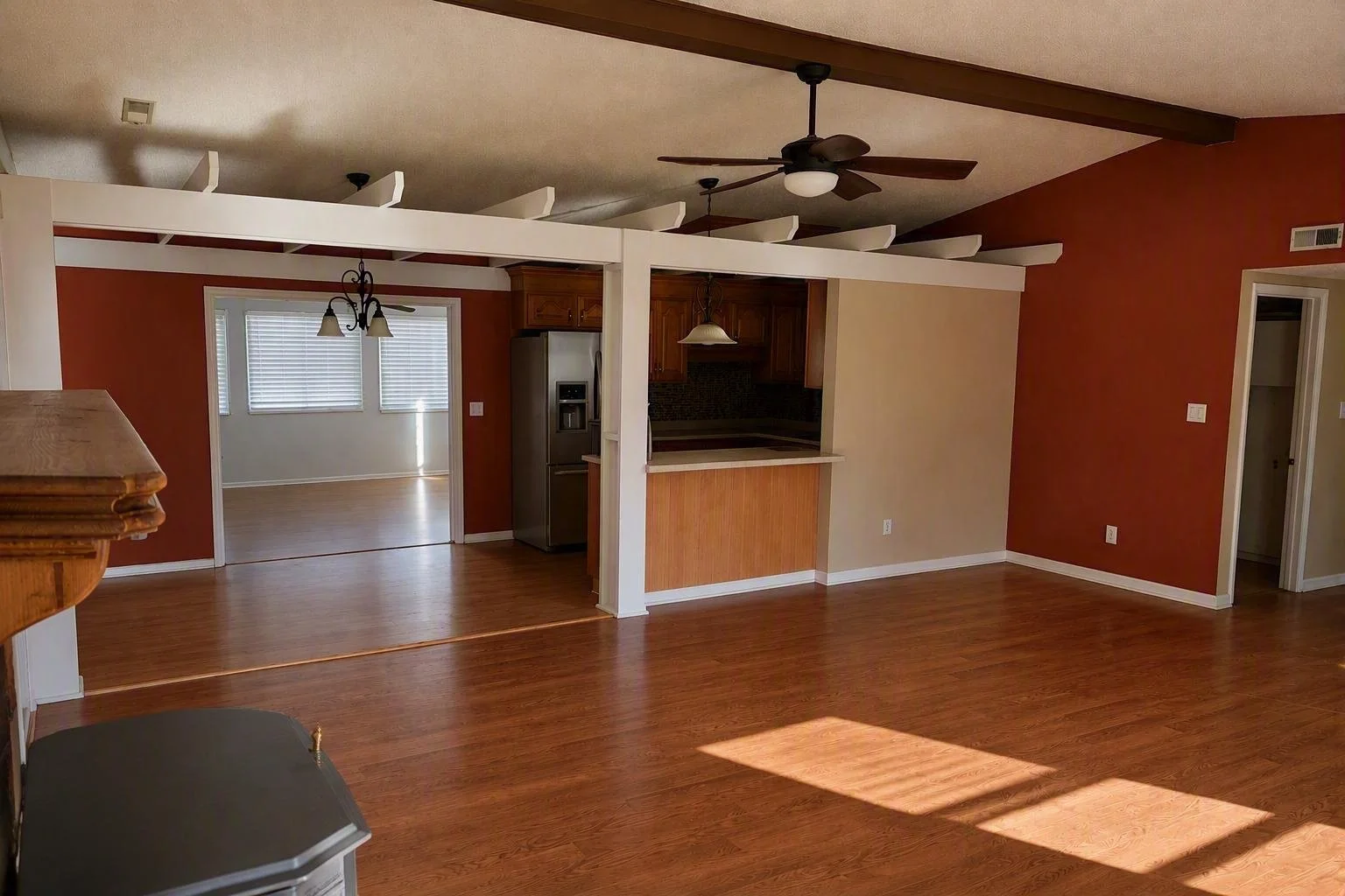Empty living room with hardwood floors, open kitchen with bar counter, ceiling fans, and a transition to a dining area with windows and blinds.