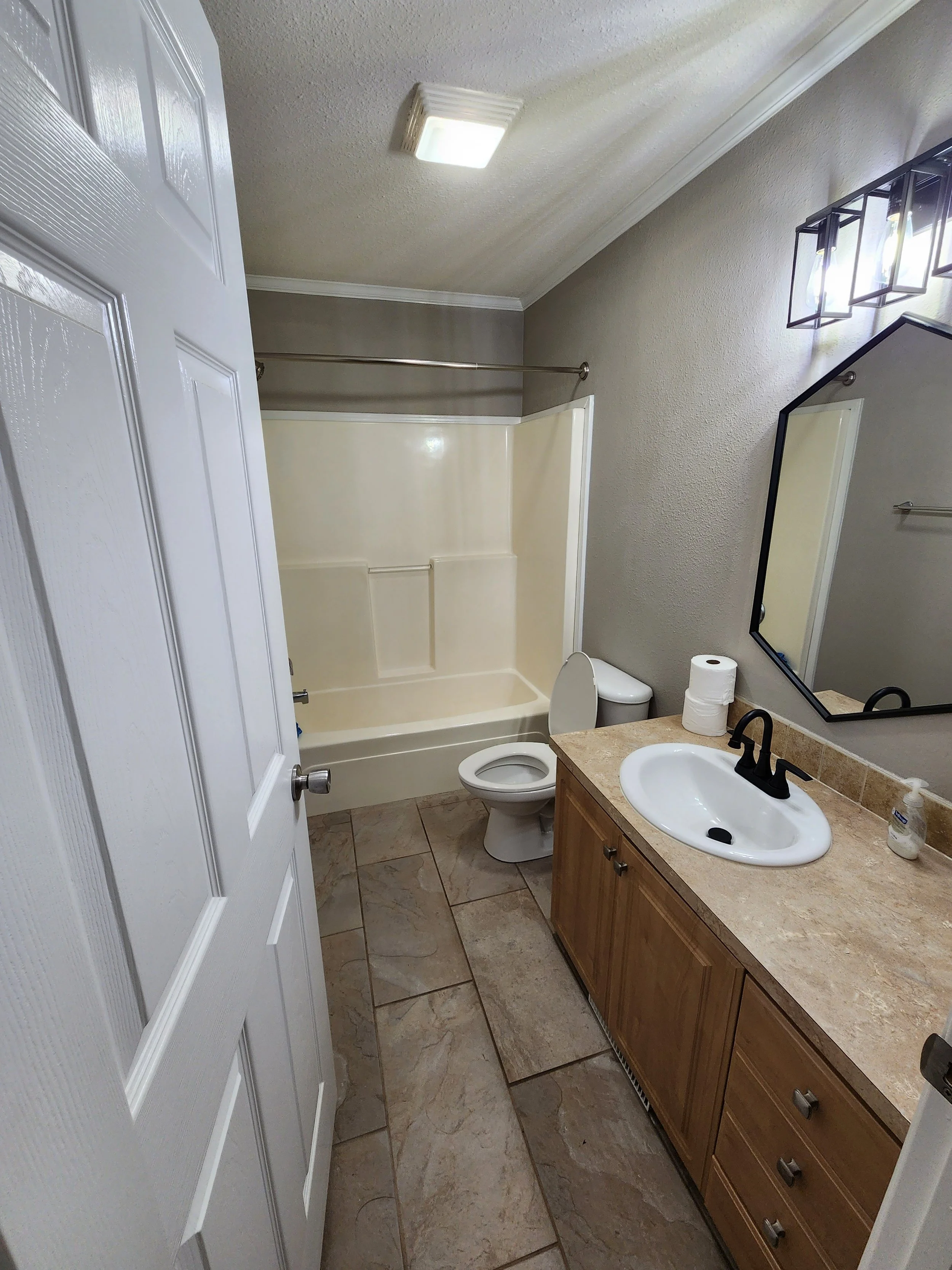 A bathroom with a white bathtub, a toilet, a wooden vanity with a sink, a mirror, a paper towel roll, and a tiled floor.