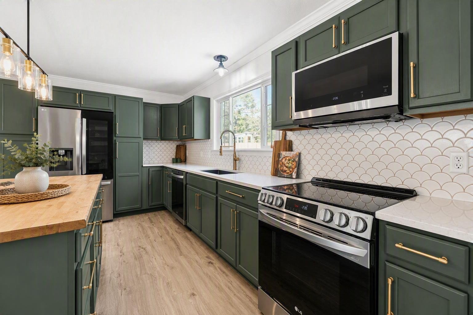 Modern kitchen with green cabinets, white countertops, stainless steel appliances, and a window above the sink. Decorative backsplash and wooden flooring.