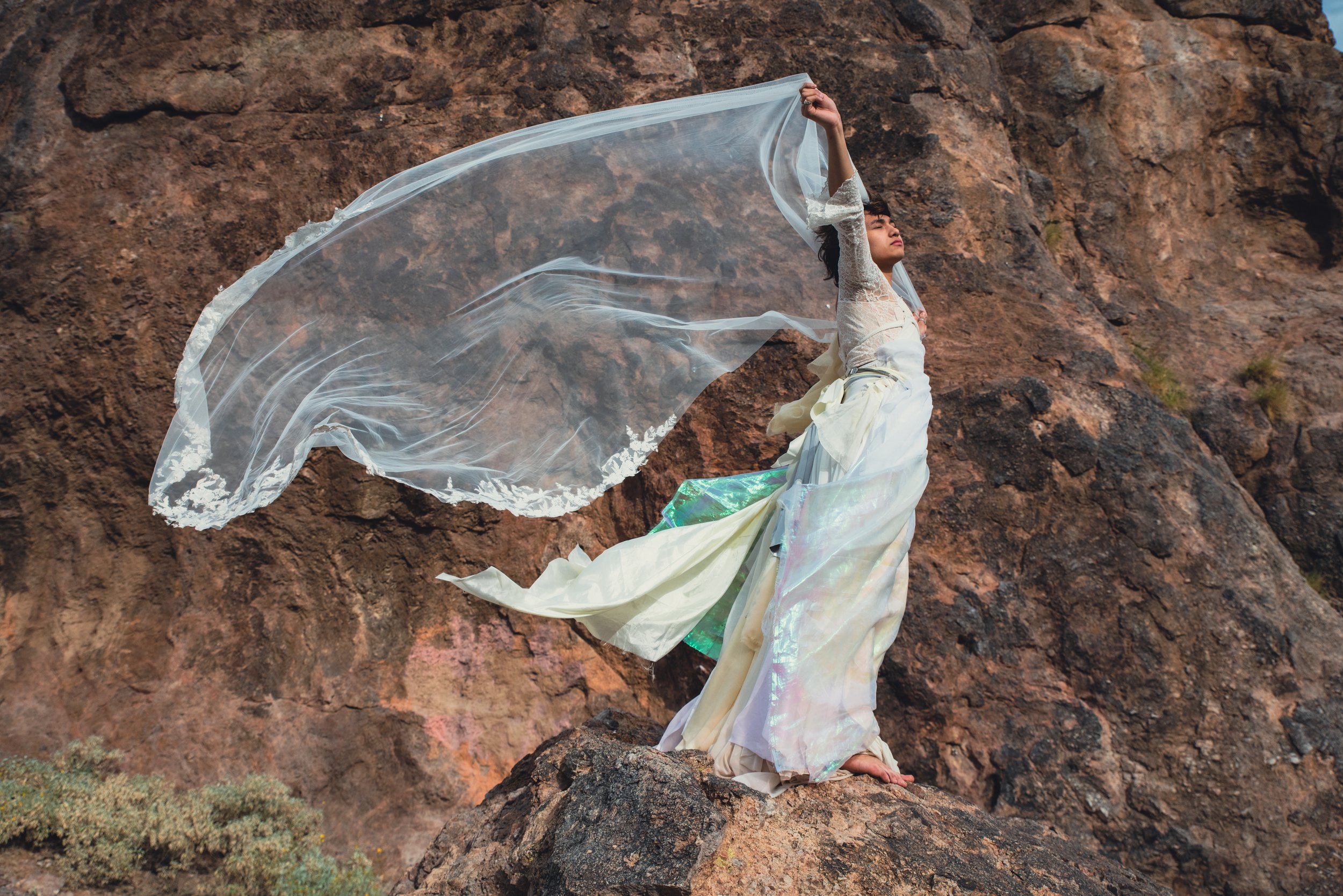 A person dressed in white with a flowing, iridescent skirt sitting on a rock in front of a rocky cliff, holding a sheer, flowing fabric above their head.