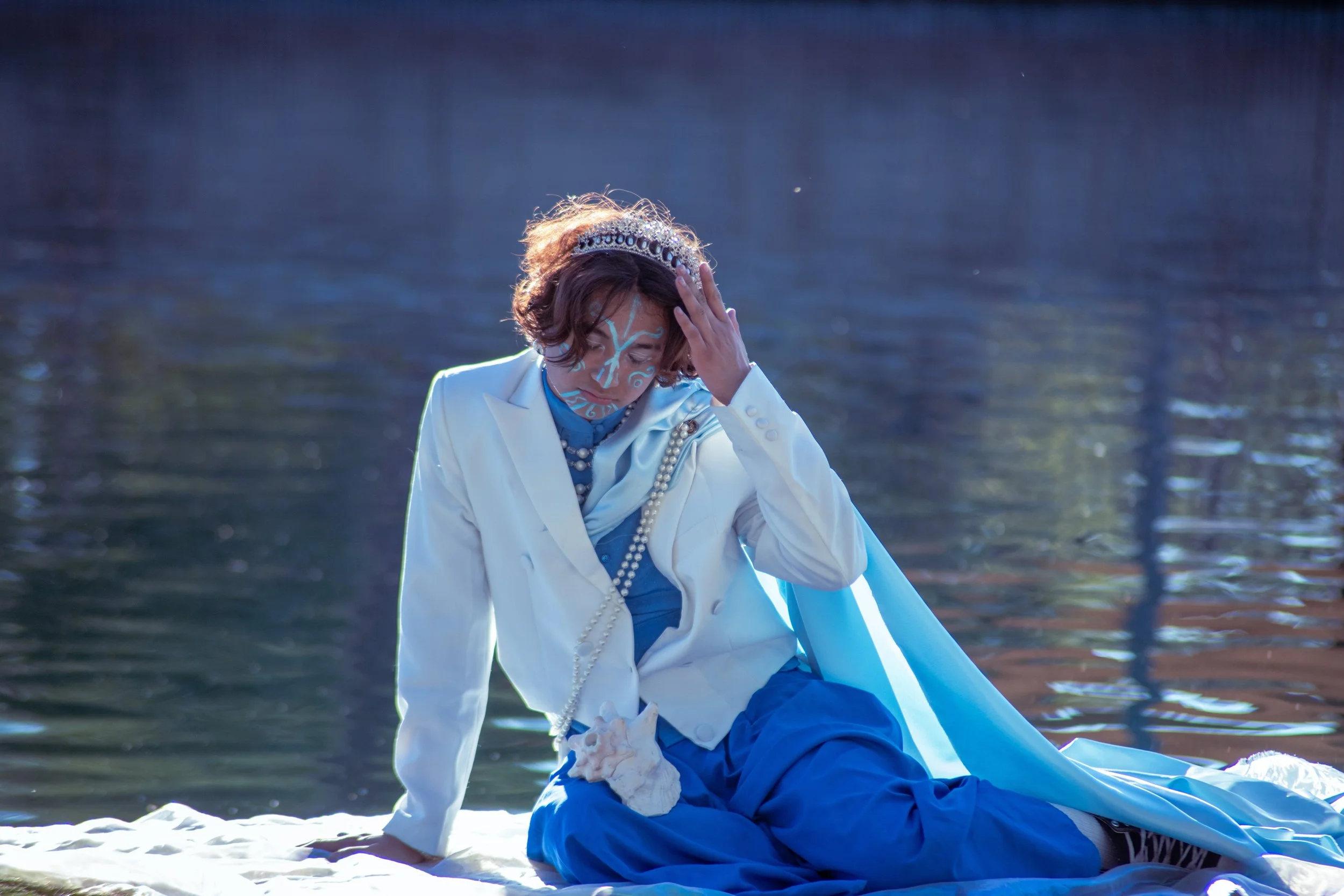 A person dressed as a royal with a tiara, face paint, pearl necklace, white blazer, and blue skirt, sitting by a body of water with a distressed expression, holding their head with one hand.