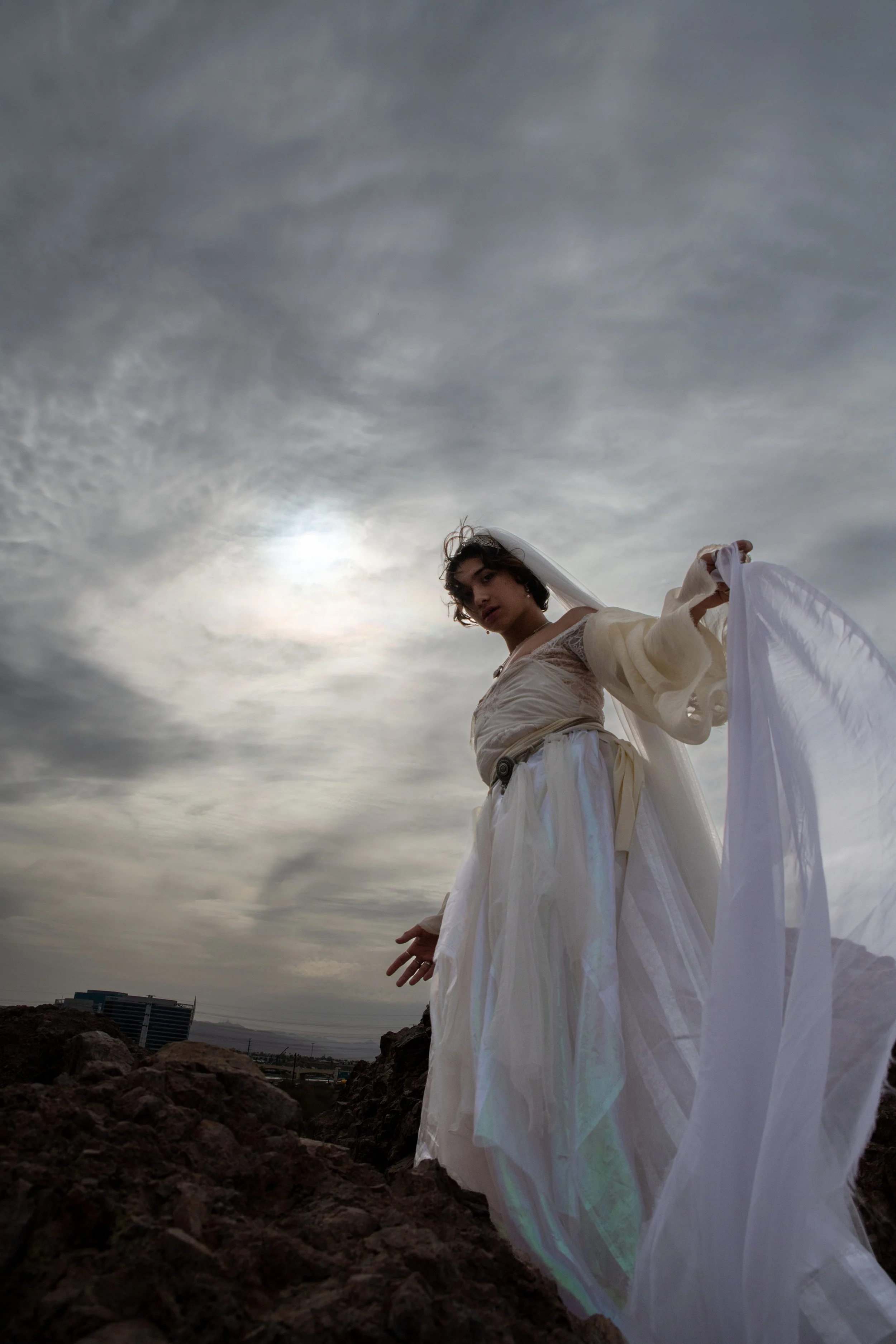 A person dressed in flowing white garments stands outdoors on a rocky terrain under an overcast sky. The person has short dark hair and is looking at the camera. The background includes a distant cityscape and skyscrapers.