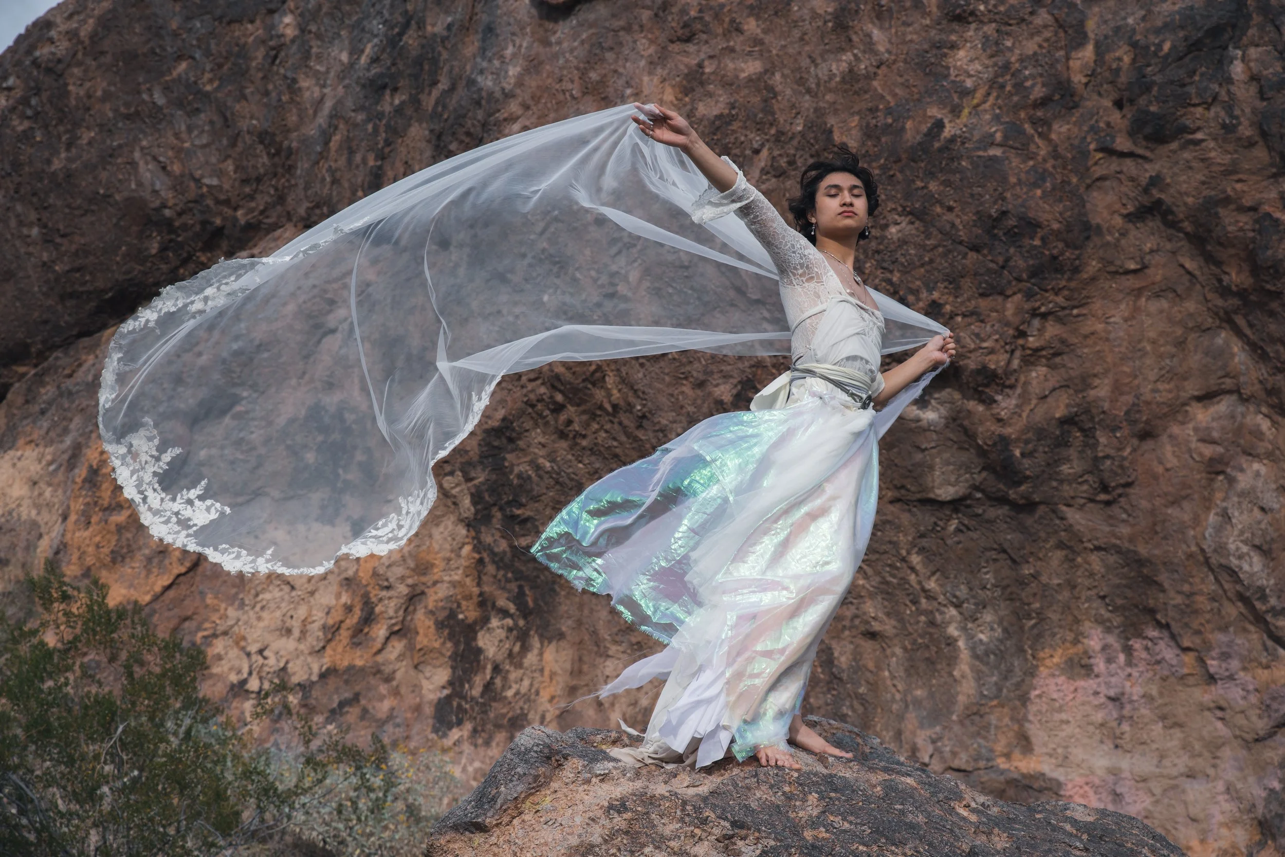A man in a white lace top and flowing iridescent skirt standing on a rock in front of a rocky cliff, holding a sheer veil that billows in the wind.