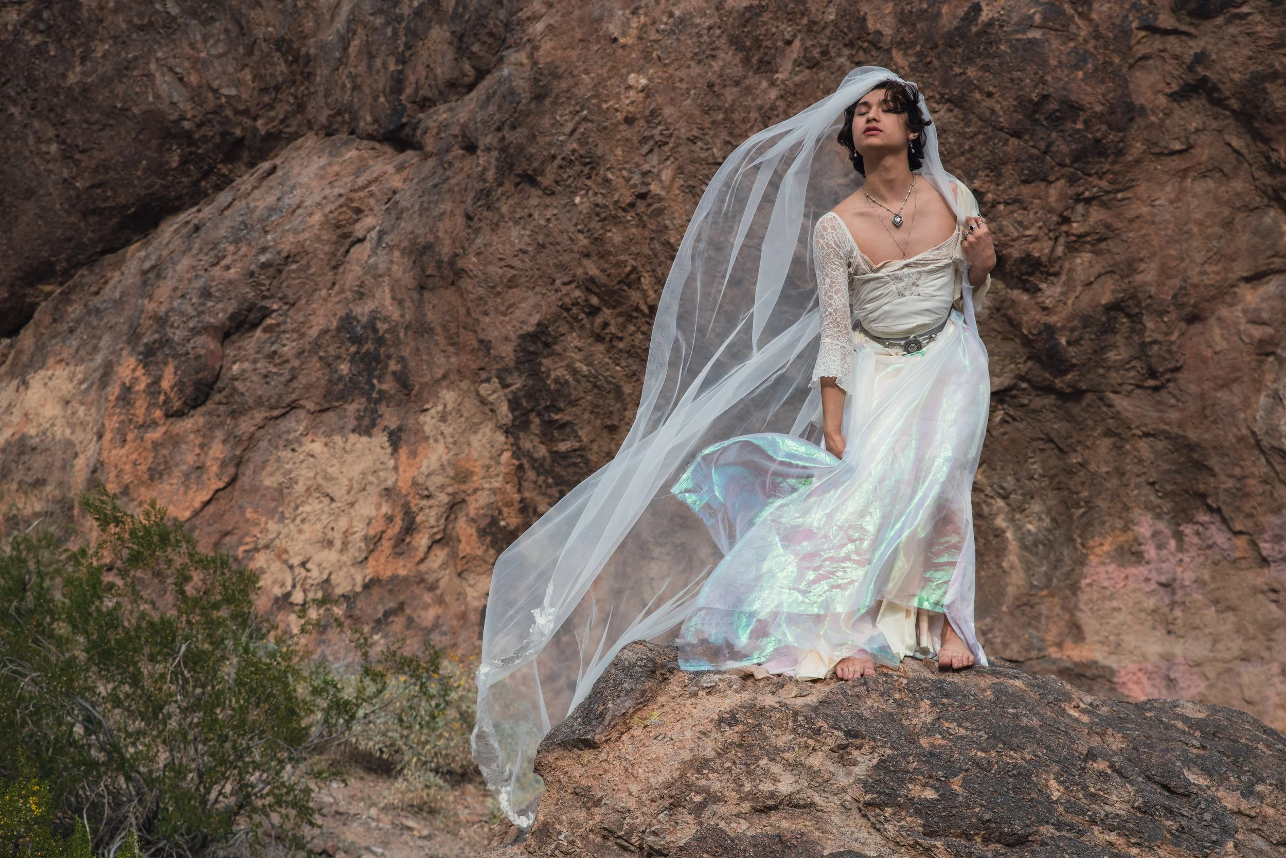 A person standing on a rock in front of a rocky mountain, dressed in a white, flowing, iridescent dress with a lace top and a veil, with jewelry including necklaces and rings, posing with eyes closed.