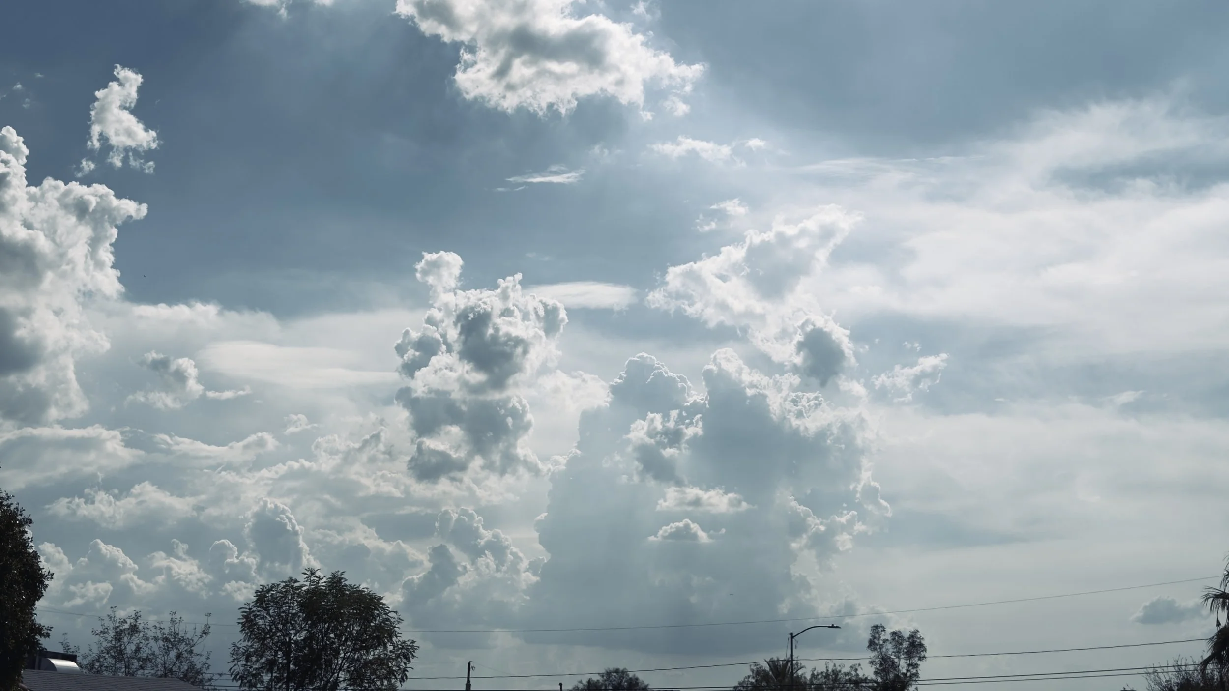 Cloudy sky with various shapes of white clouds and visible blue sky, trees and rooftops in the foreground.
