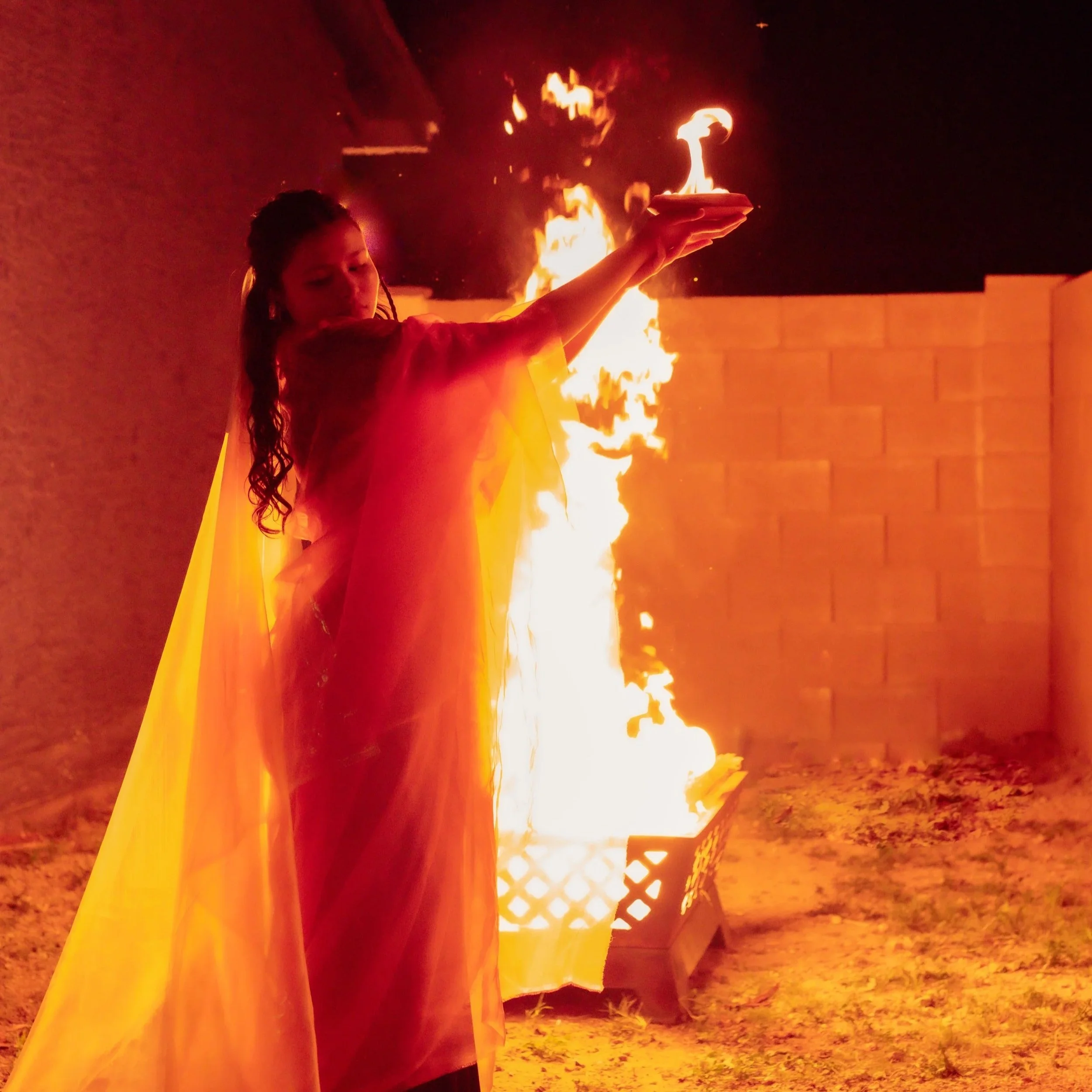 A young woman with wet, dark hair holding a plate with a flaming dish, standing in front of a brick wall with flames rising behind her.