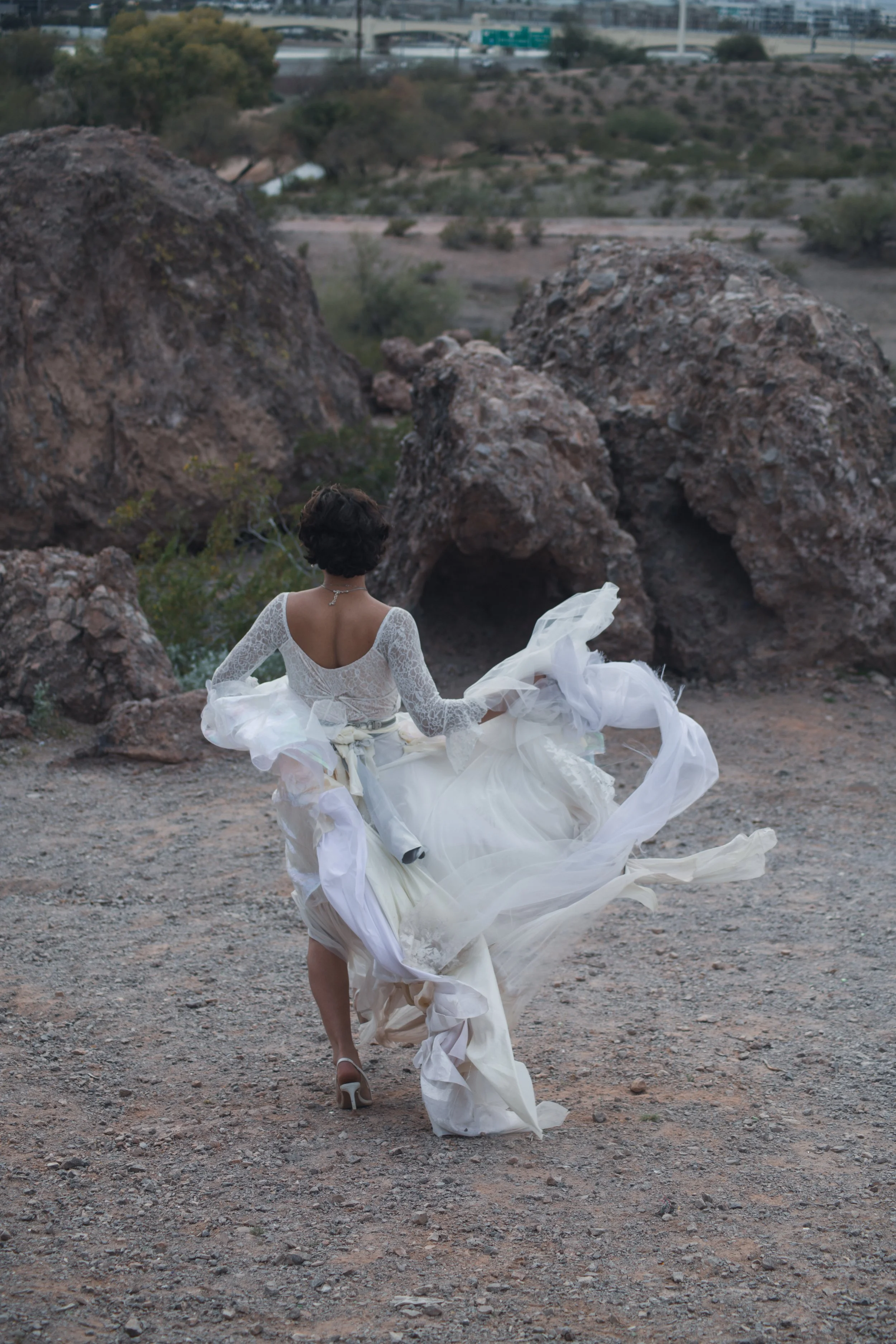 Woman in a white wedding dress walking through rocky desert landscape, with volcanic rock formations in the background.