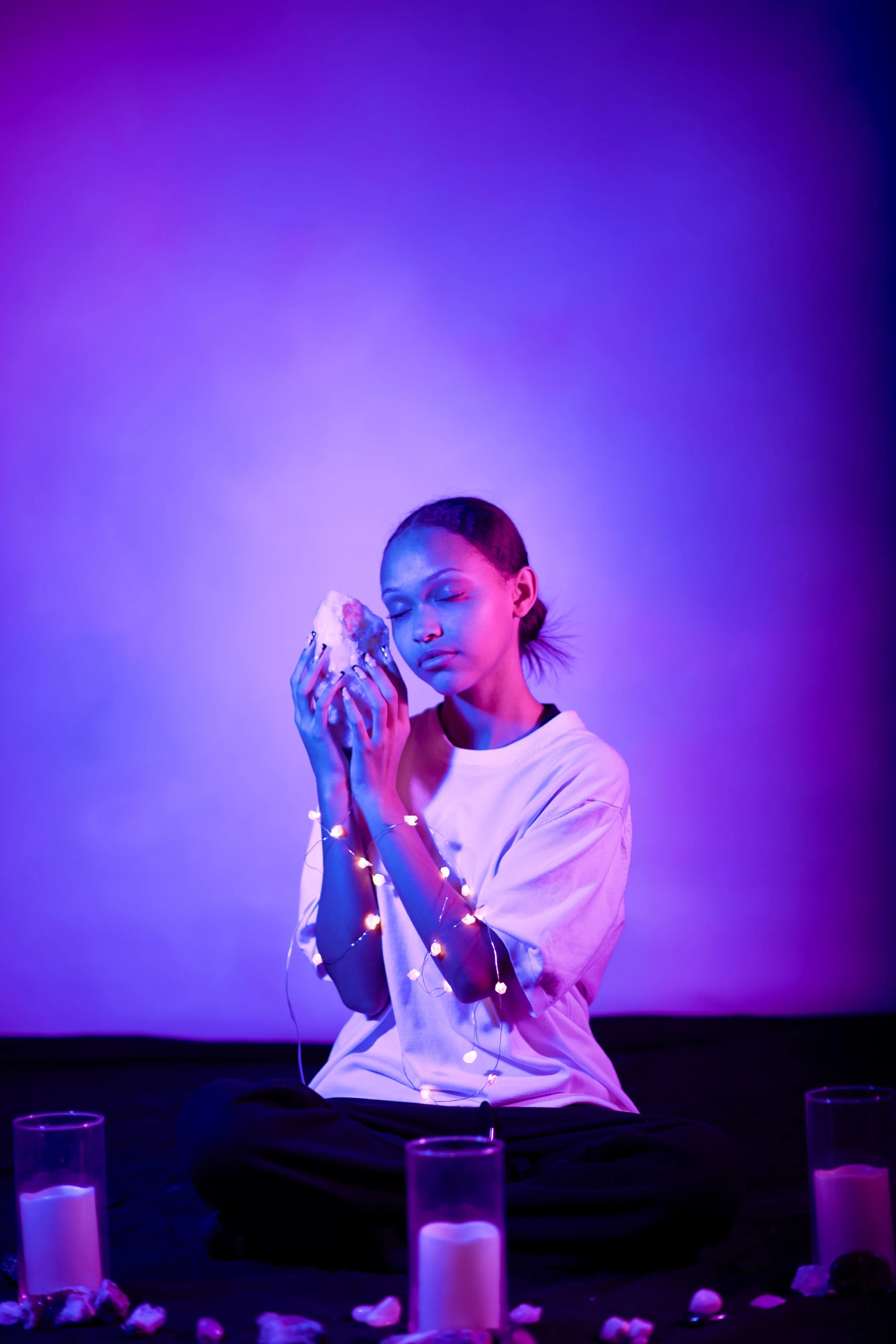 A woman sitting cross-legged at night is holding a large crystal with her eyes closed. She is surrounded by candles and flower petals, and decorated with string lights on her arms. The background is illuminated with purple and blue lighting.