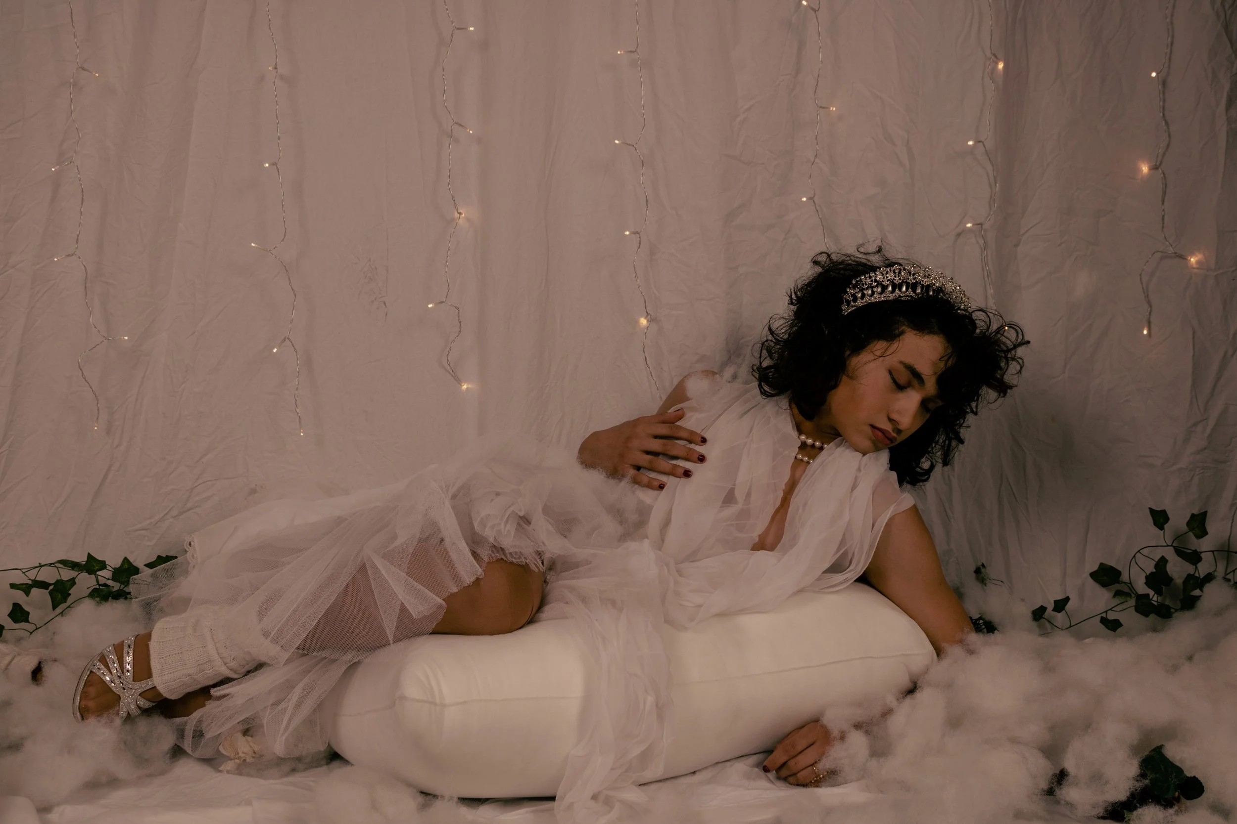 A woman dressed in white lying on a bed with a backdrop of string lights and greenery, wearing a tiara and jewelry, with her eyes closed and an elegant pose.