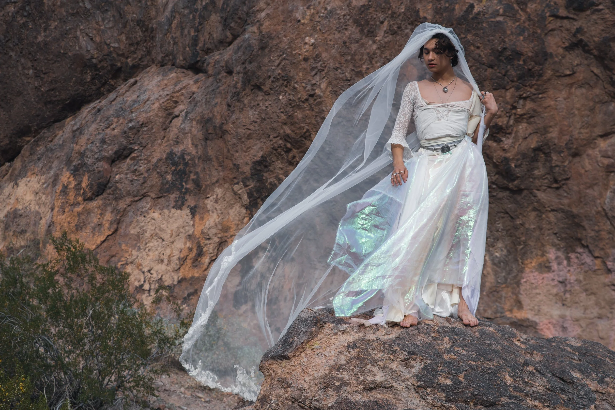A man in a wedding dress and veil standing barefoot on a rocky cliff with a desert landscape background.