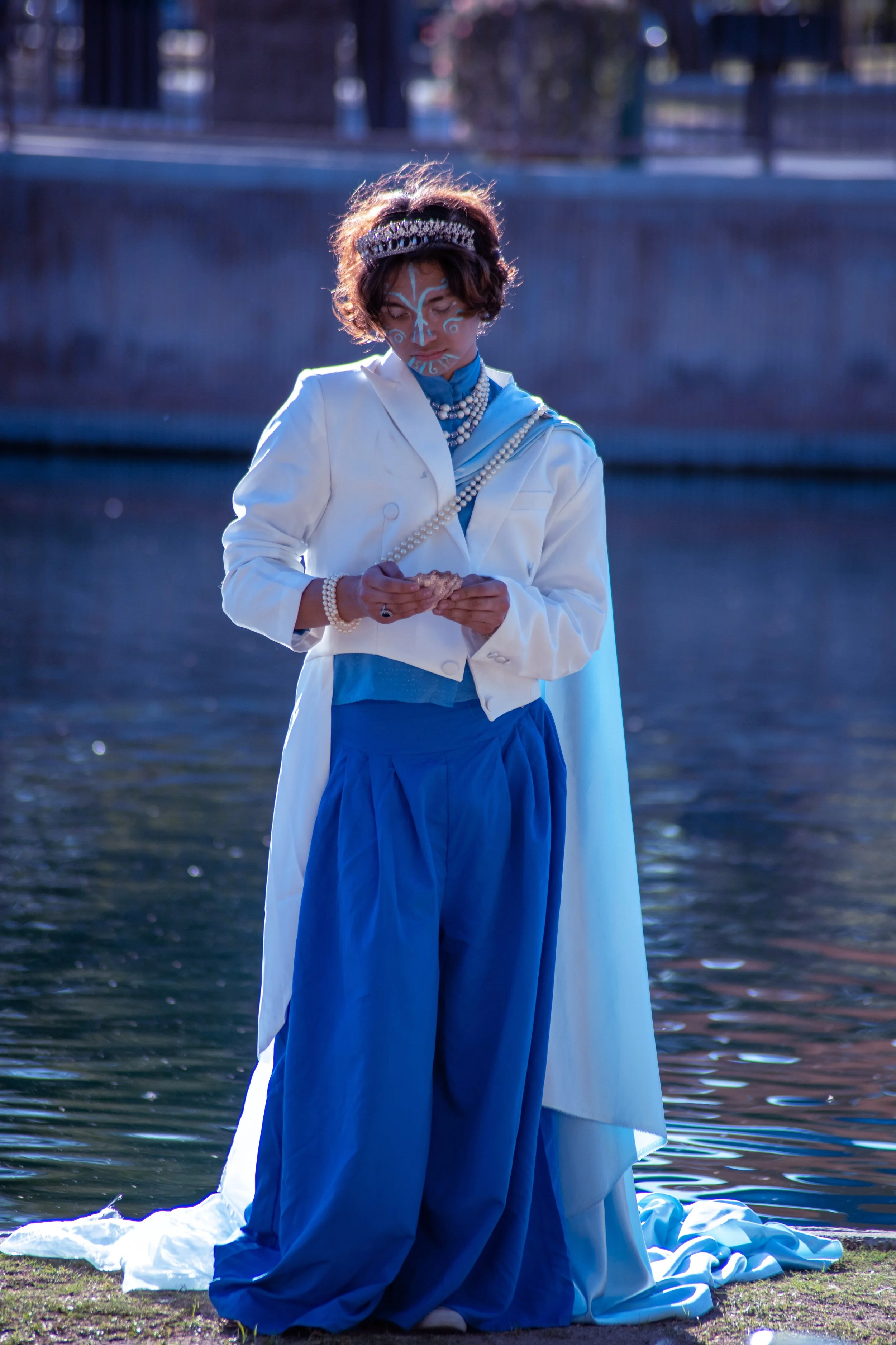 Man in a white jacket and blue dress with face paint, wearing a crown and pearl necklaces, kneeling by a body of water, holding a shell.