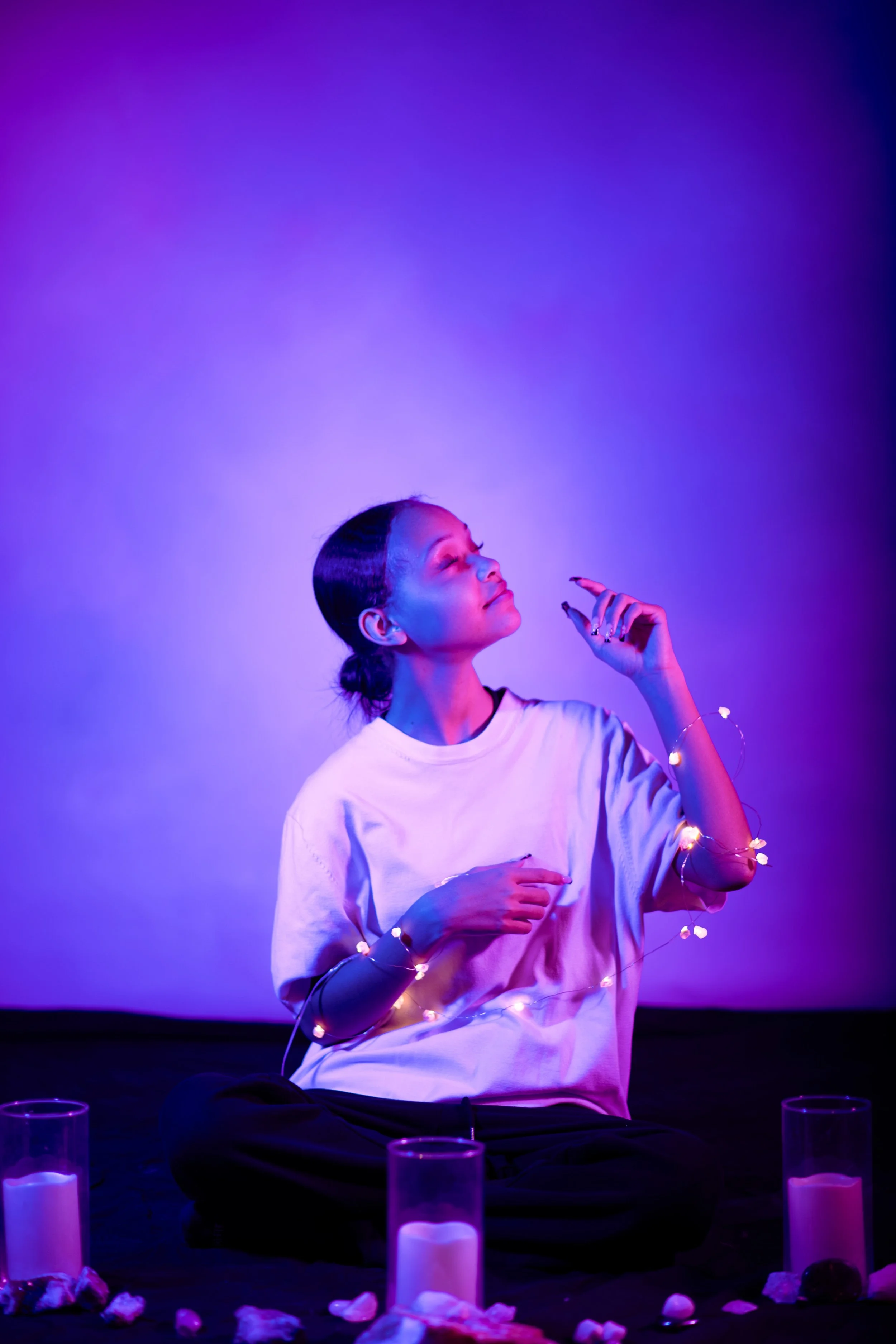 A woman with dark hair tied back, sitting cross-legged, surrounded by glowing candles and rose petals, holding a string of fairy lights, illuminated by purple and blue lighting.